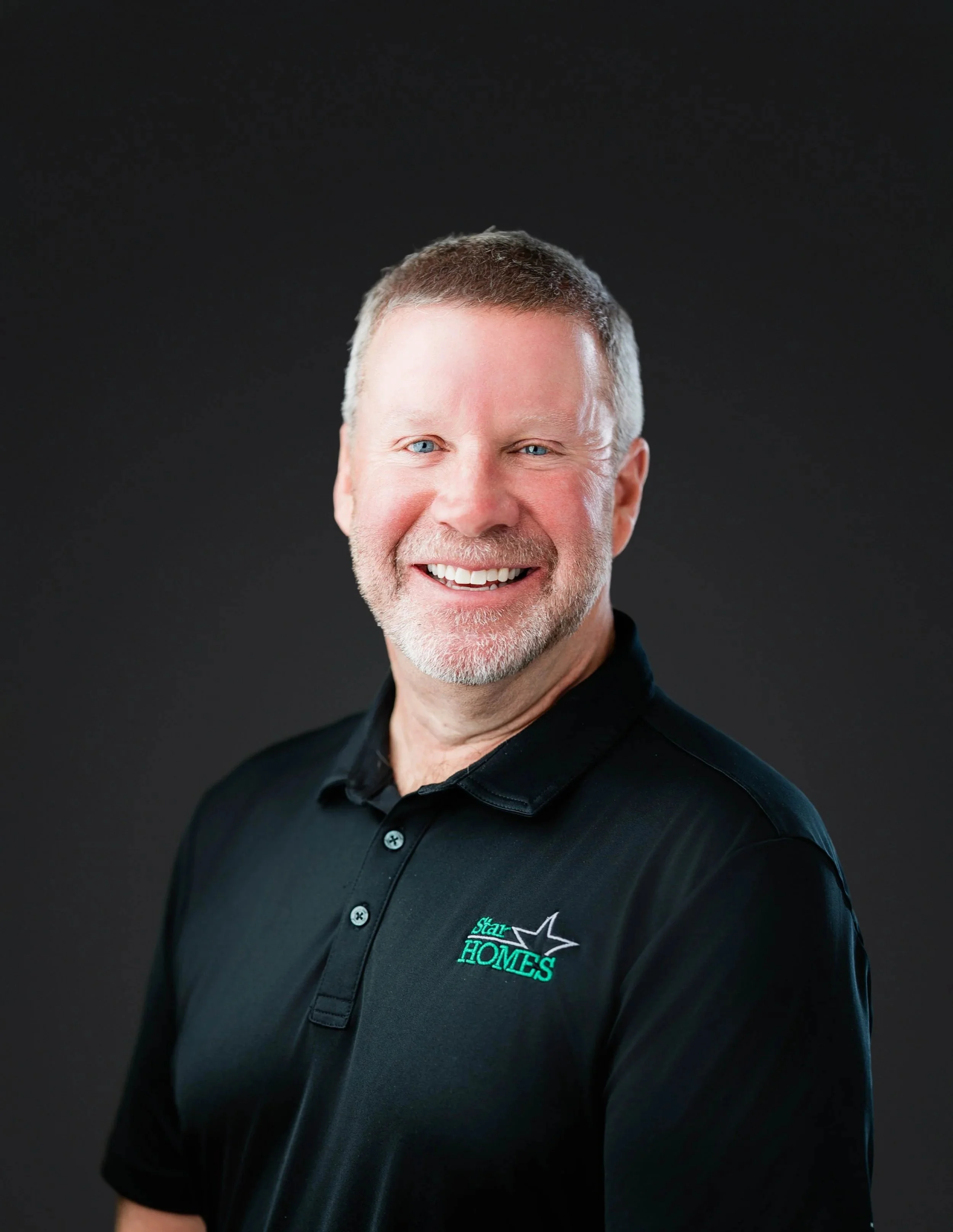 Close-up of a smiling middle-aged man with short gray hair and blue eyes, wearing a black collared shirt with an embroidered logo that reads 'Star Homes' with a star, set against a dark background.