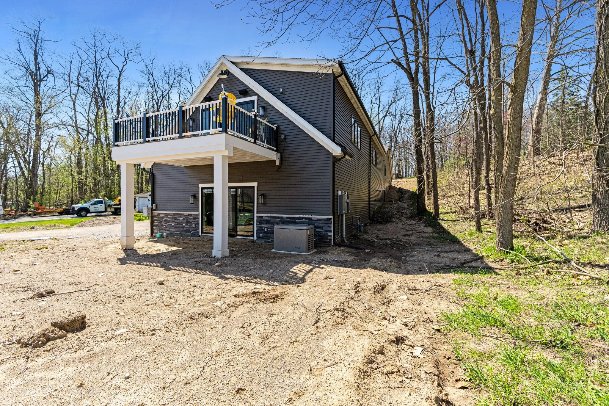 Newly constructed two-story house with gray siding, a rear balcony with black railing, and a support pillar beneath it, surrounded by a wooded area with some dirt and grass on the ground.