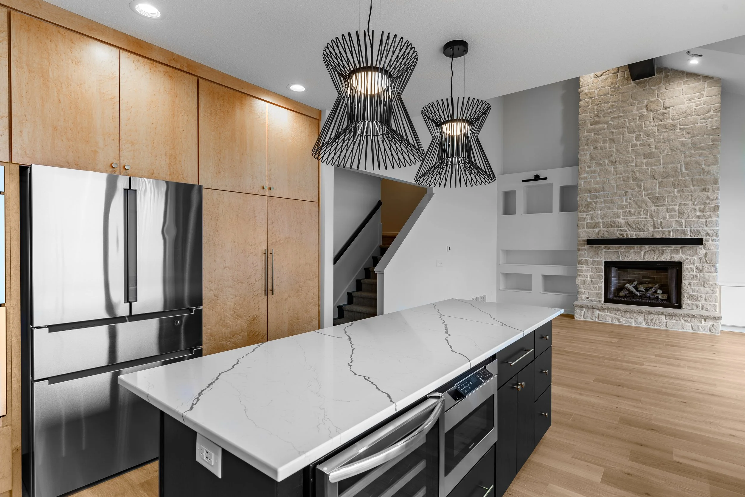 Modern kitchen with black and white island, stainless steel refrigerator, wood cabinets, and a stone fireplace in the background.