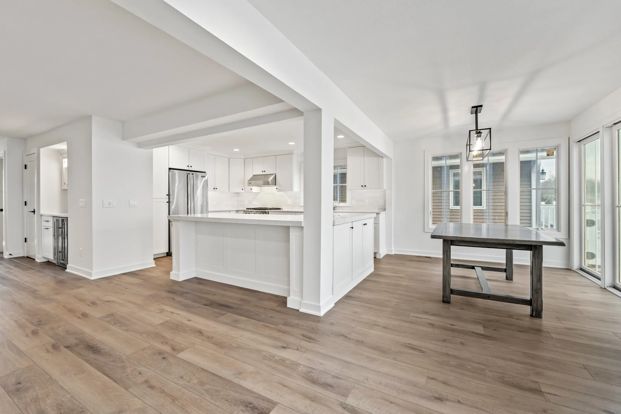 Modern kitchen with white cabinetry and stainless steel appliances, adjacent to a dining area with a rustic wooden table, large windows, and hardwood flooring.
