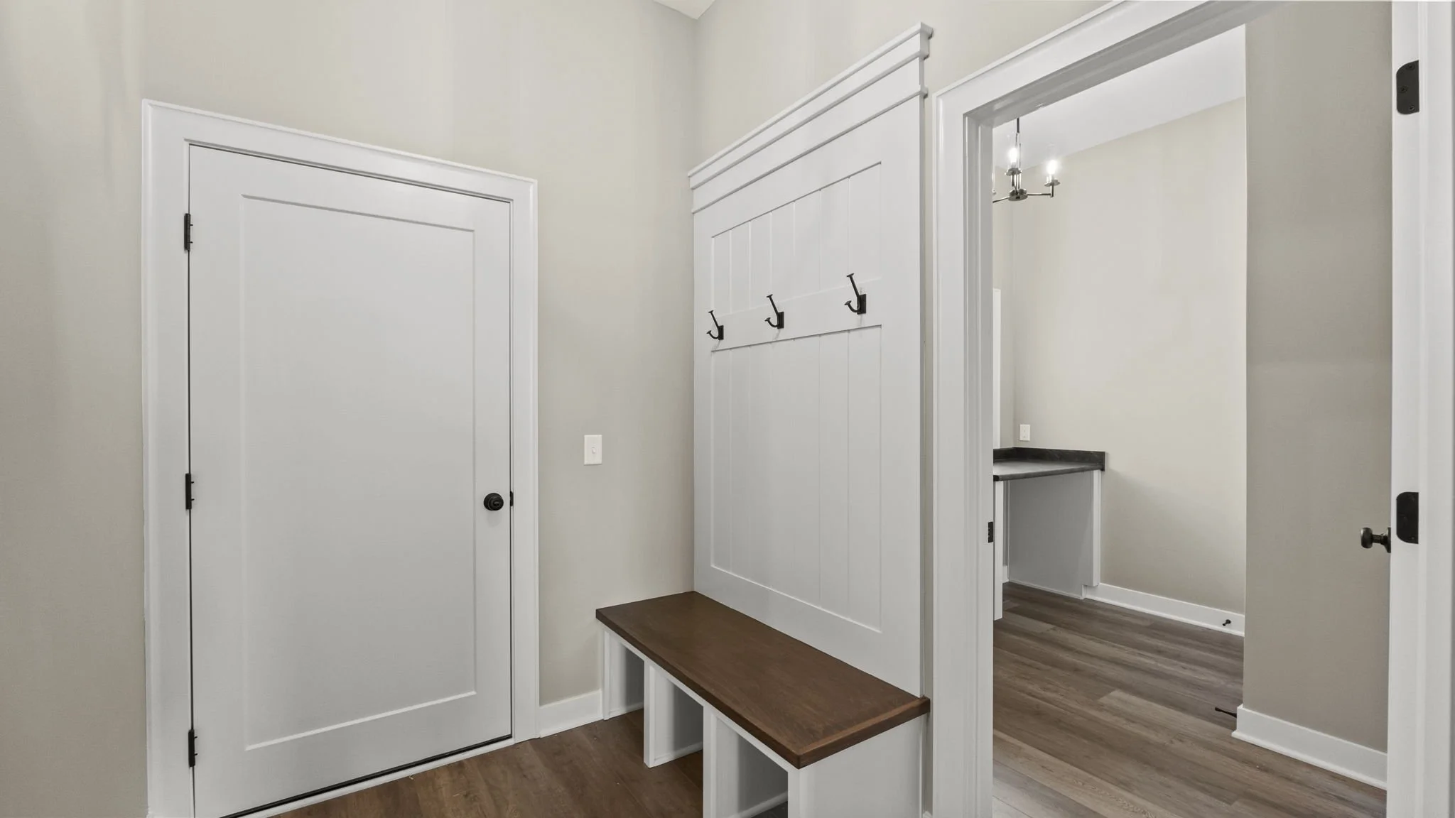 Entrance hallway with a white door, wall hooks, a wooden bench, and a view into a room with a small countertop and a chandelier.