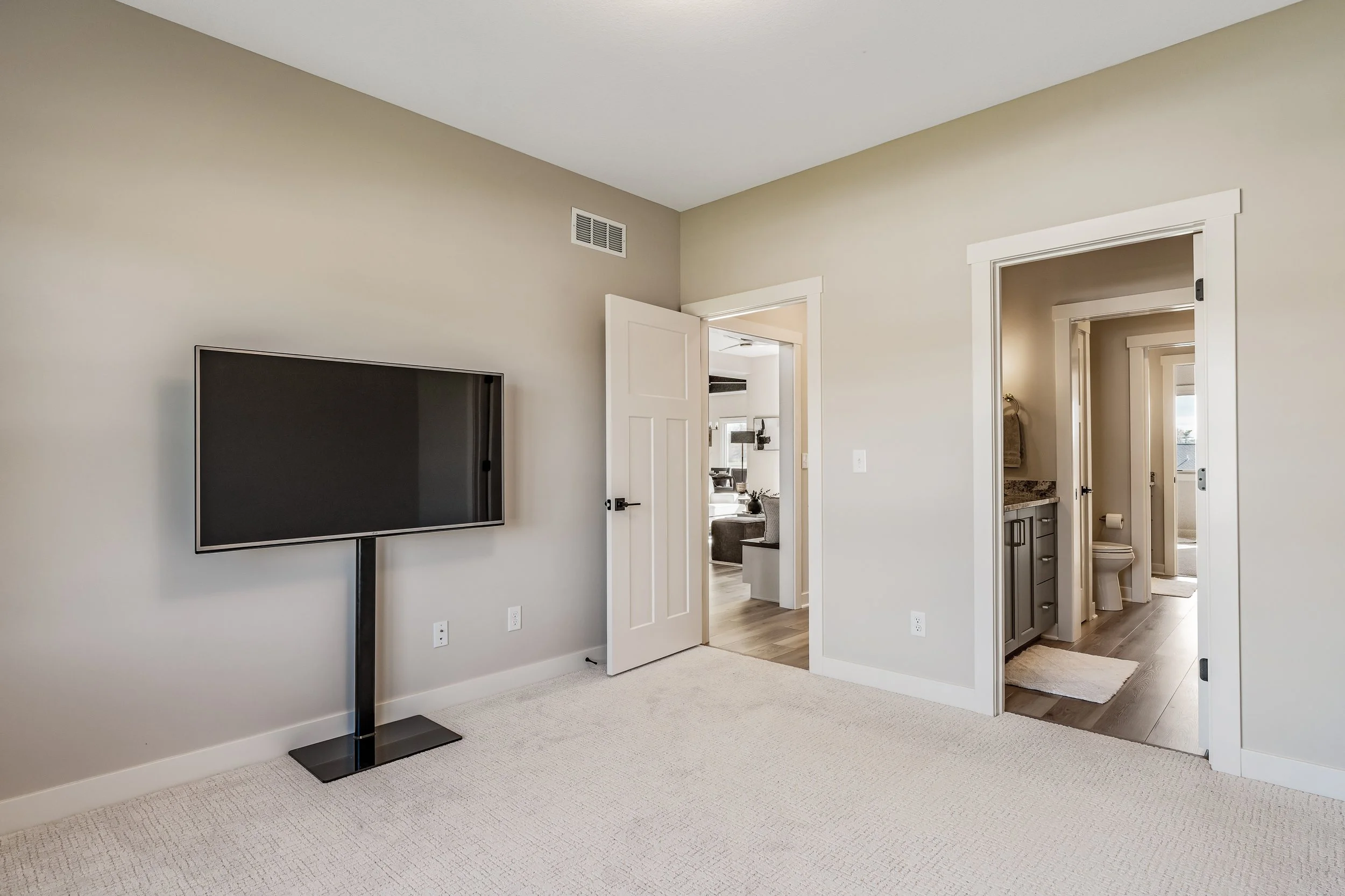 Empty living room with a wall-mounted flat-screen TV, beige walls, doors leading to a bedroom, bathroom, and another room, light-colored carpet, and a remote control on the TV stand.