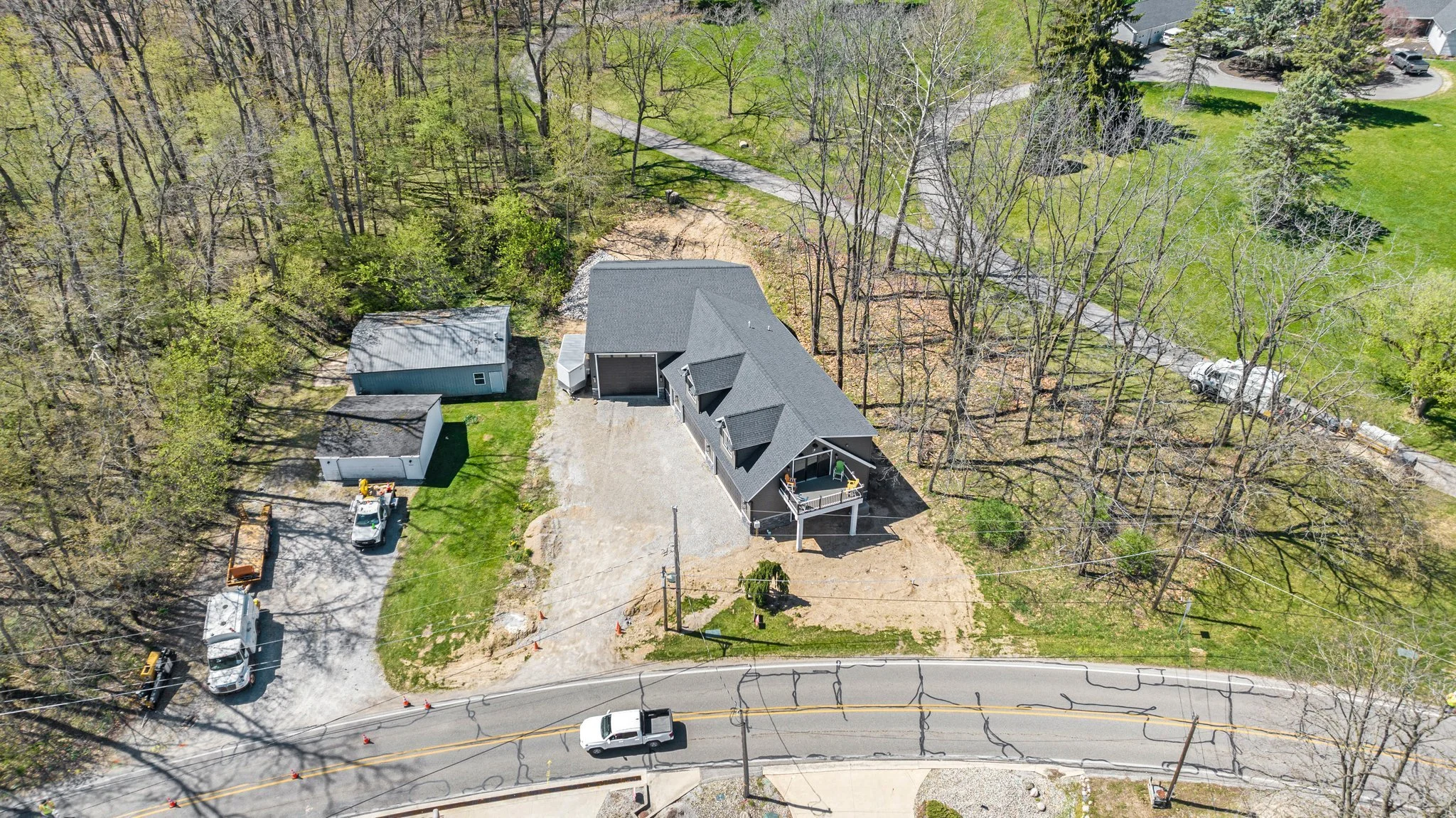 An aerial view of a house under construction, with a large driveway, a couple of outbuildings, surrounded by trees, with a curved road in front and a landscaped lawn.