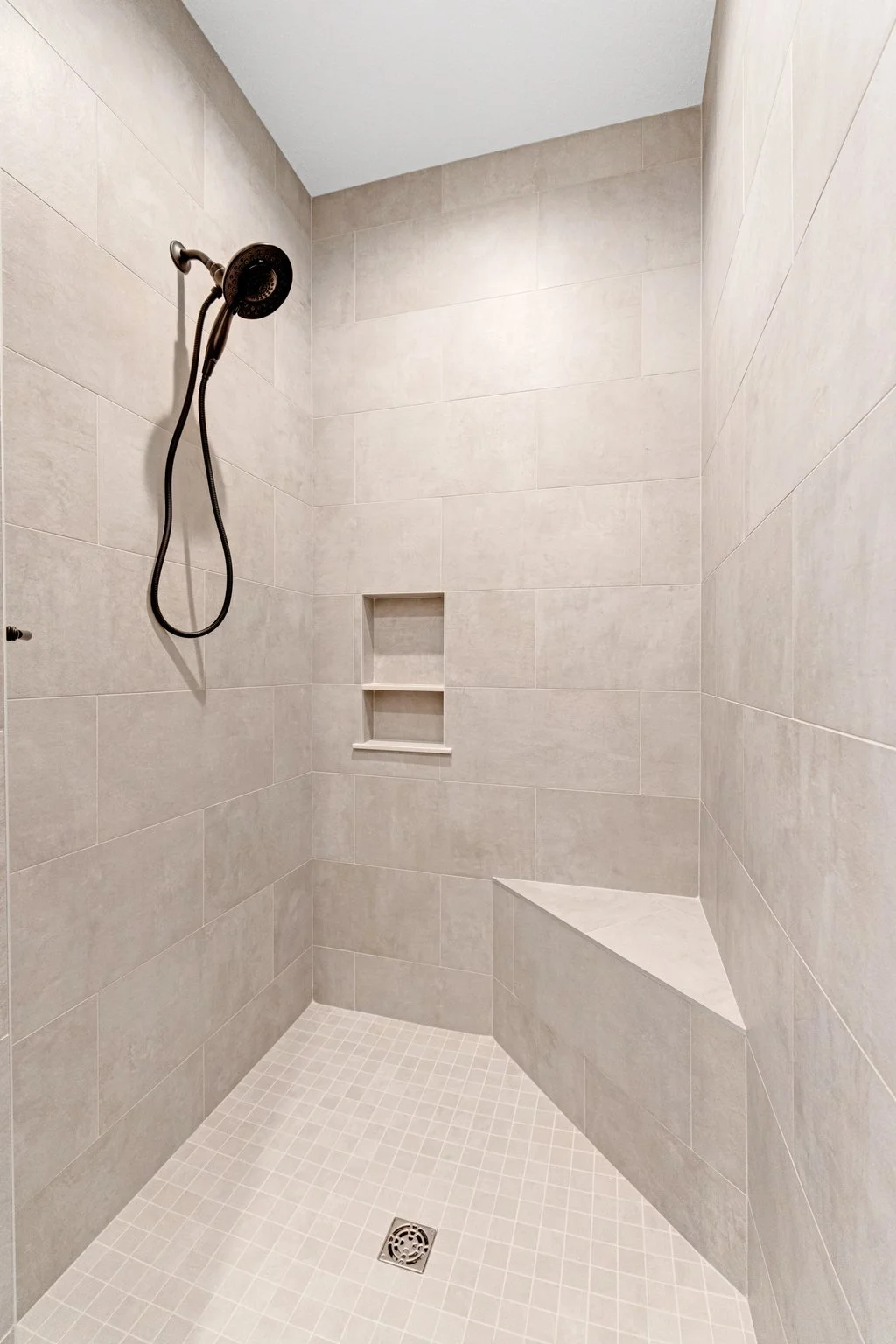 A clean shower with beige tiled walls, a built-in shelf, a corner seat, a black showerhead, and a drain on the tile floor.