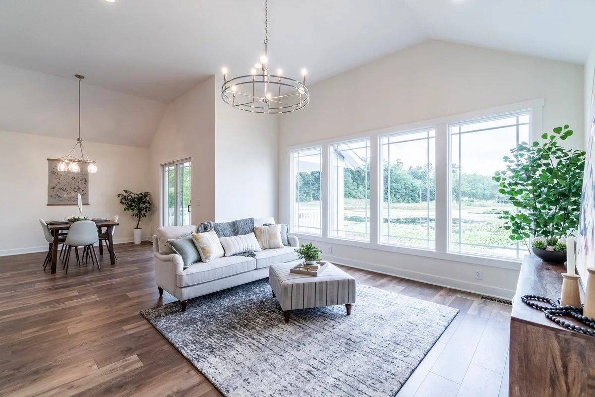 Bright living room with large windows, white walls, wooden flooring, a light-colored sofa, an armchair, a striped ottoman, and a modern chandelier, with a dining area in the background and decorative plants.