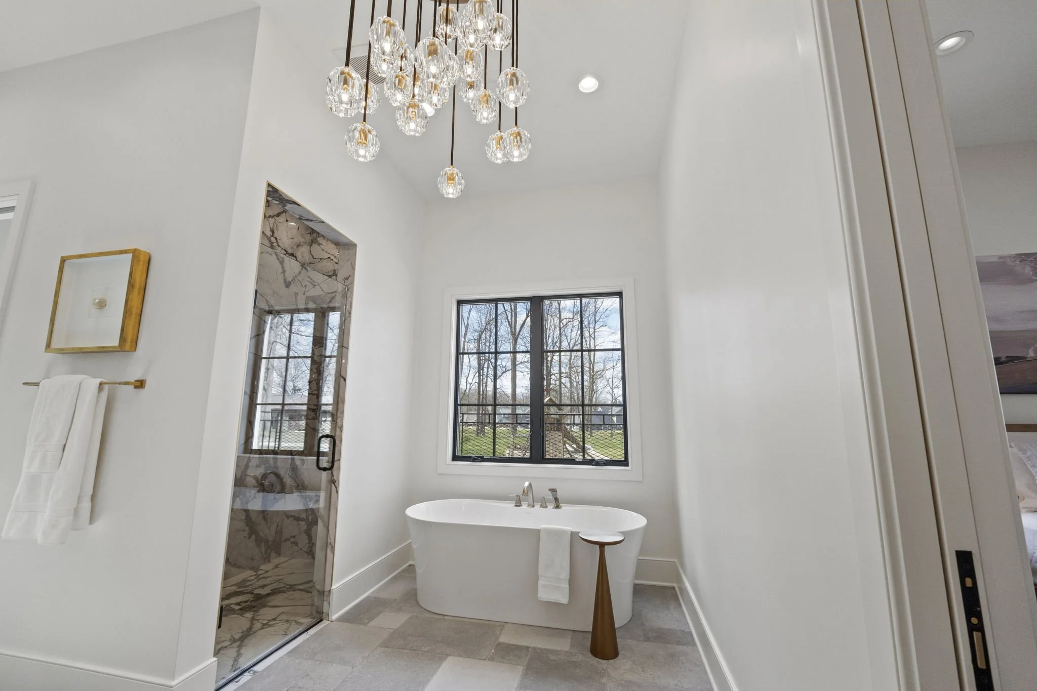Modern bathroom with a white bathtub, large window showing trees outside, marble shower, gold-framed mirror, and modern chandelier.