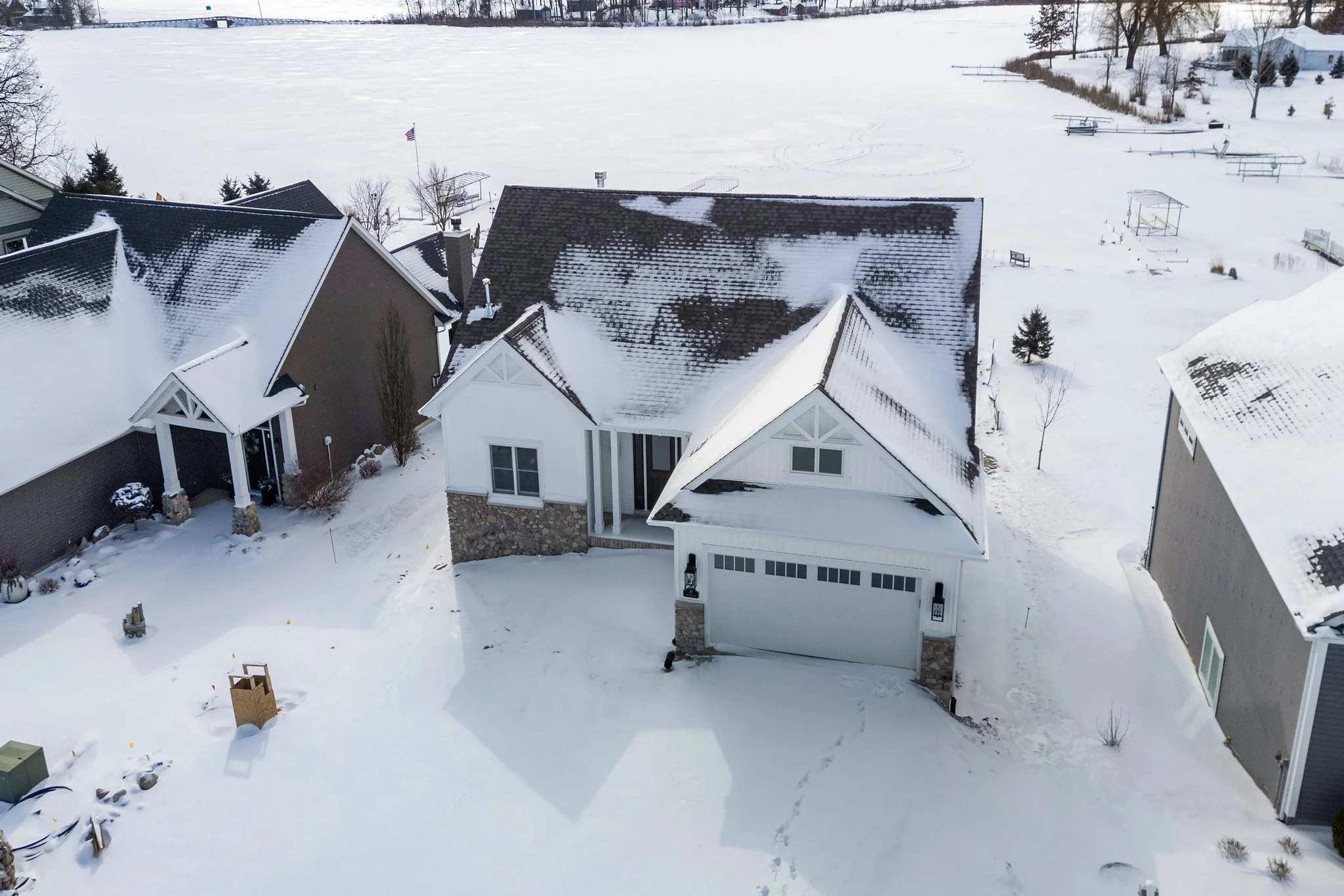 Aerial view of a snow-covered suburban house with a white facade, stone accents, and a garage, surrounded by neighboring houses, trees, and a frozen lake in the background.