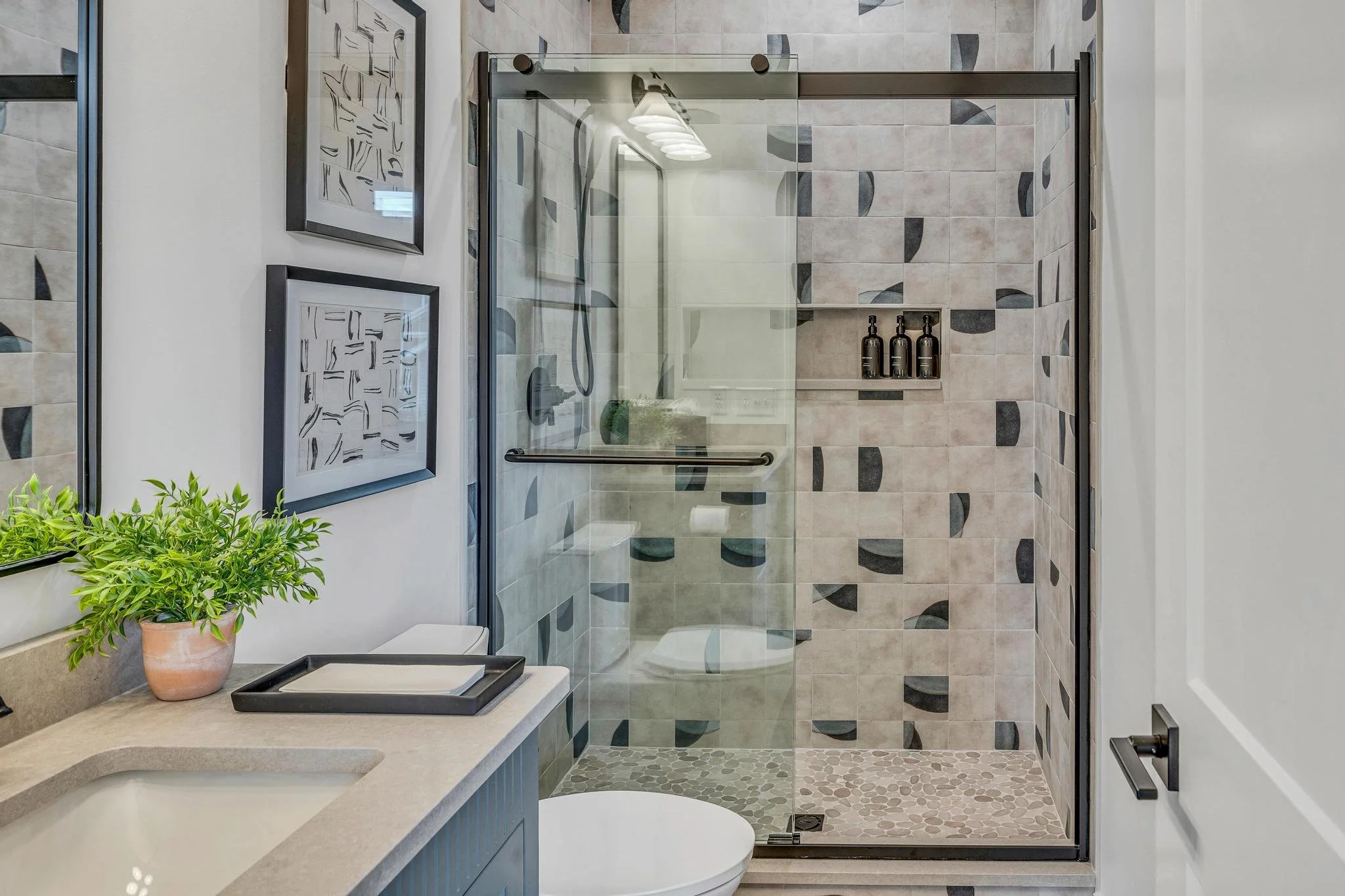 Bathroom with a glass shower enclosure, patterned tile walls, a white toilet, a small countertop with a potted plant, and framed artwork on the wall.