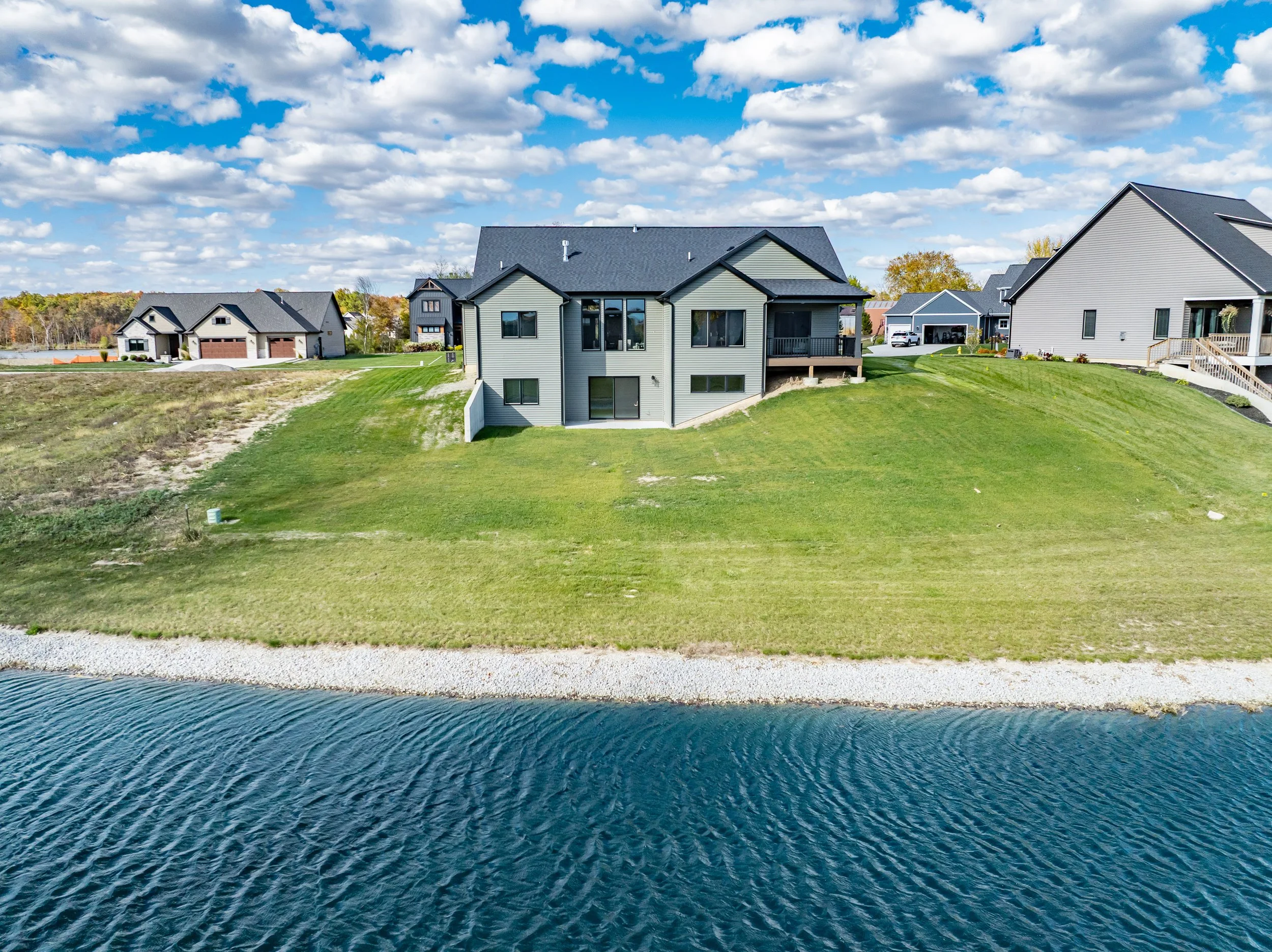 A house on a grassy hill by a body of water with neighboring houses in the background and partly cloudy sky.