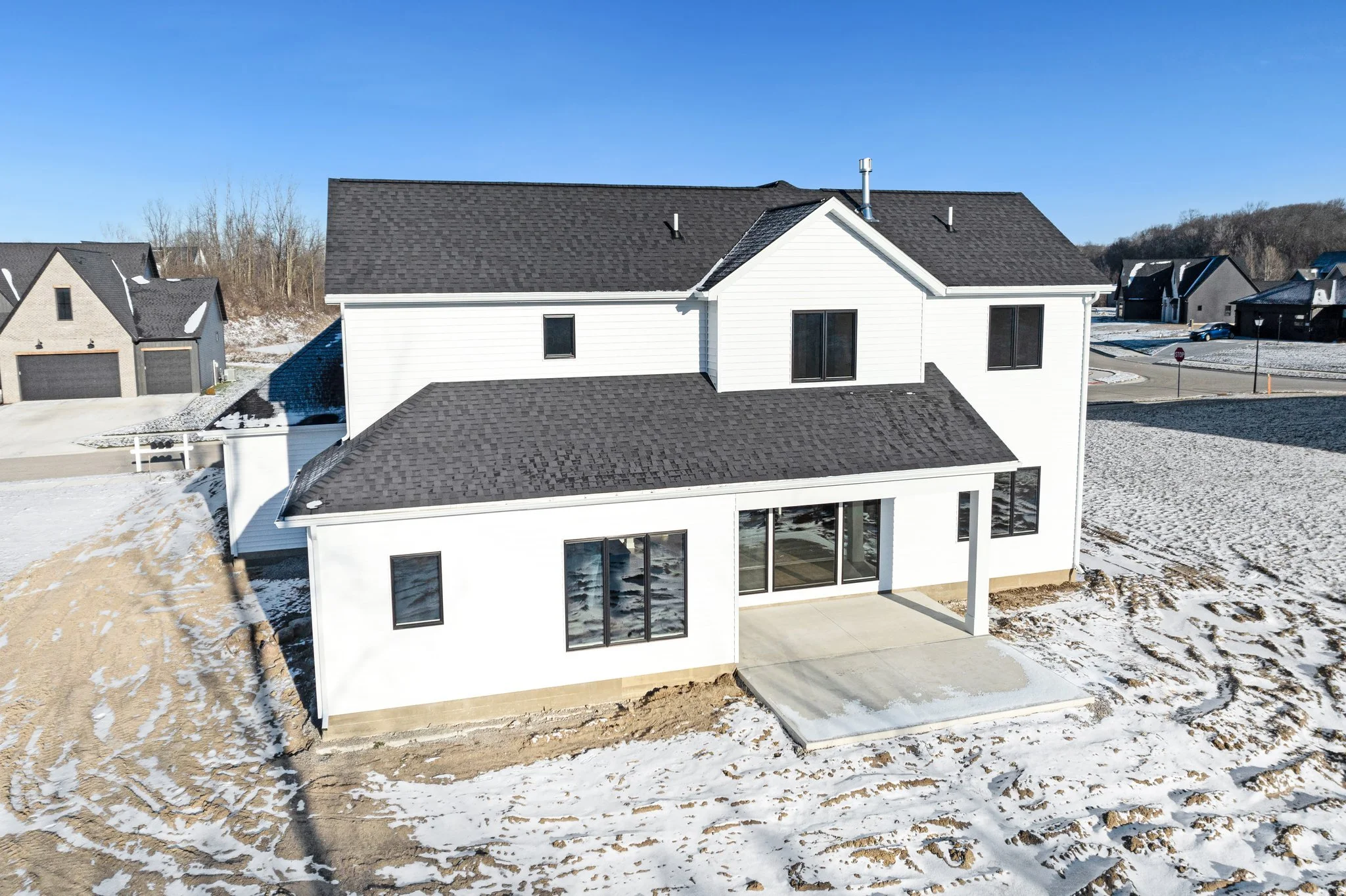 A newly constructed white house with black roof and large glass doors, situated on a snowy lot in a residential neighborhood.