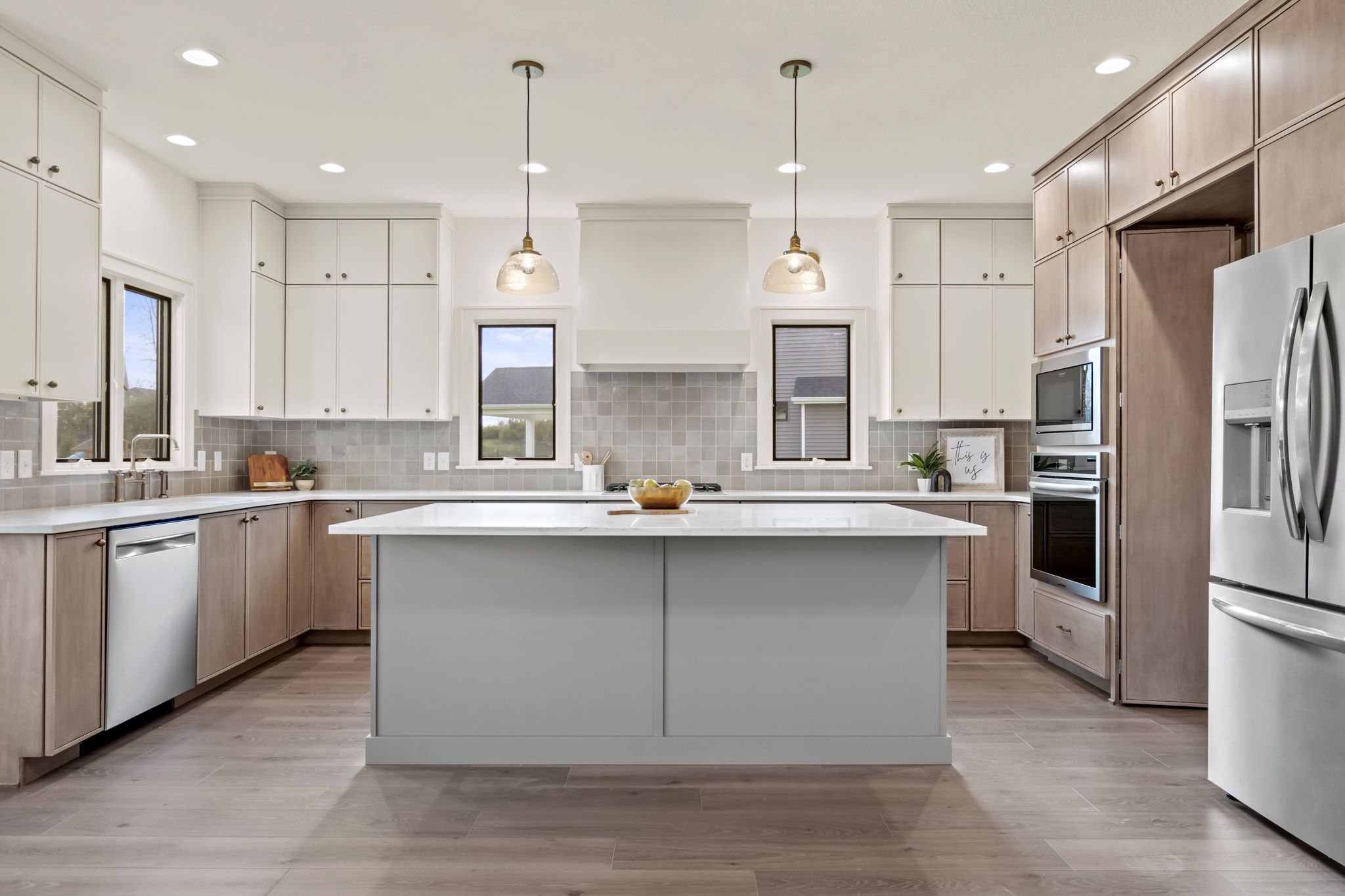 Modern kitchen with white and light wood cabinetry, a central island, stainless steel appliances, and pendant lighting, with windows allowing natural light.