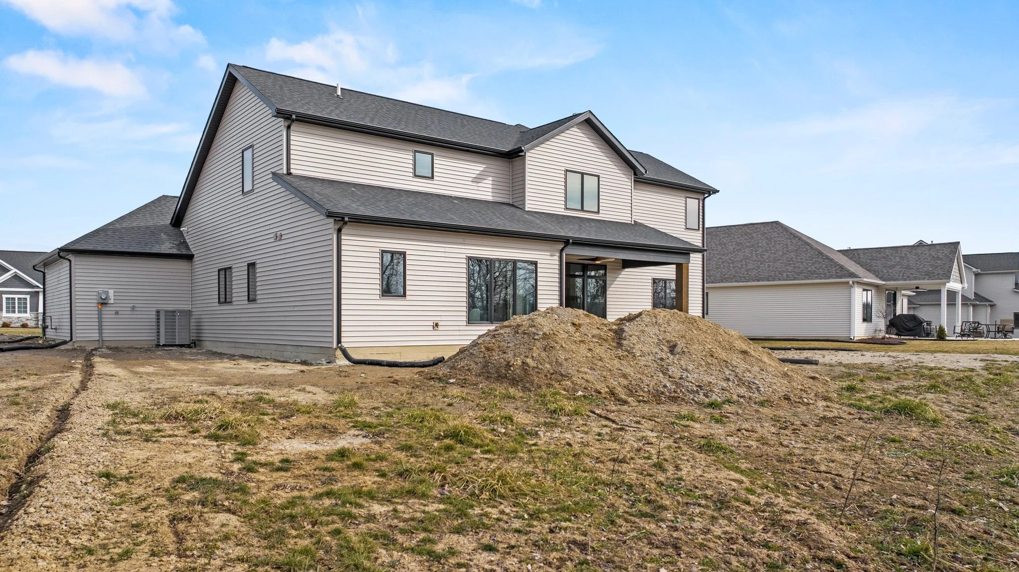 A two-story house under construction with dirt and a pile of soil in the foreground, neighboring houses in the background, and a partly cloudy sky.