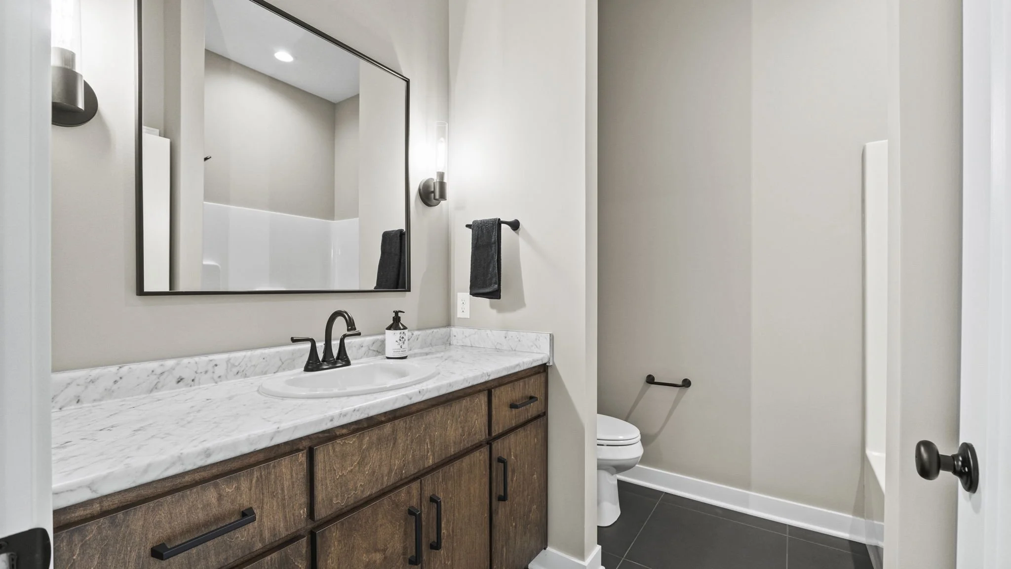 A bathroom with a wooden vanity with a white marble countertop, a black faucet, and a soap dispenser. A large mirror hangs above the vanity. A gray towel is on a rod next to the mirror. The toilet is to the right of the vanity, and a black towel is o