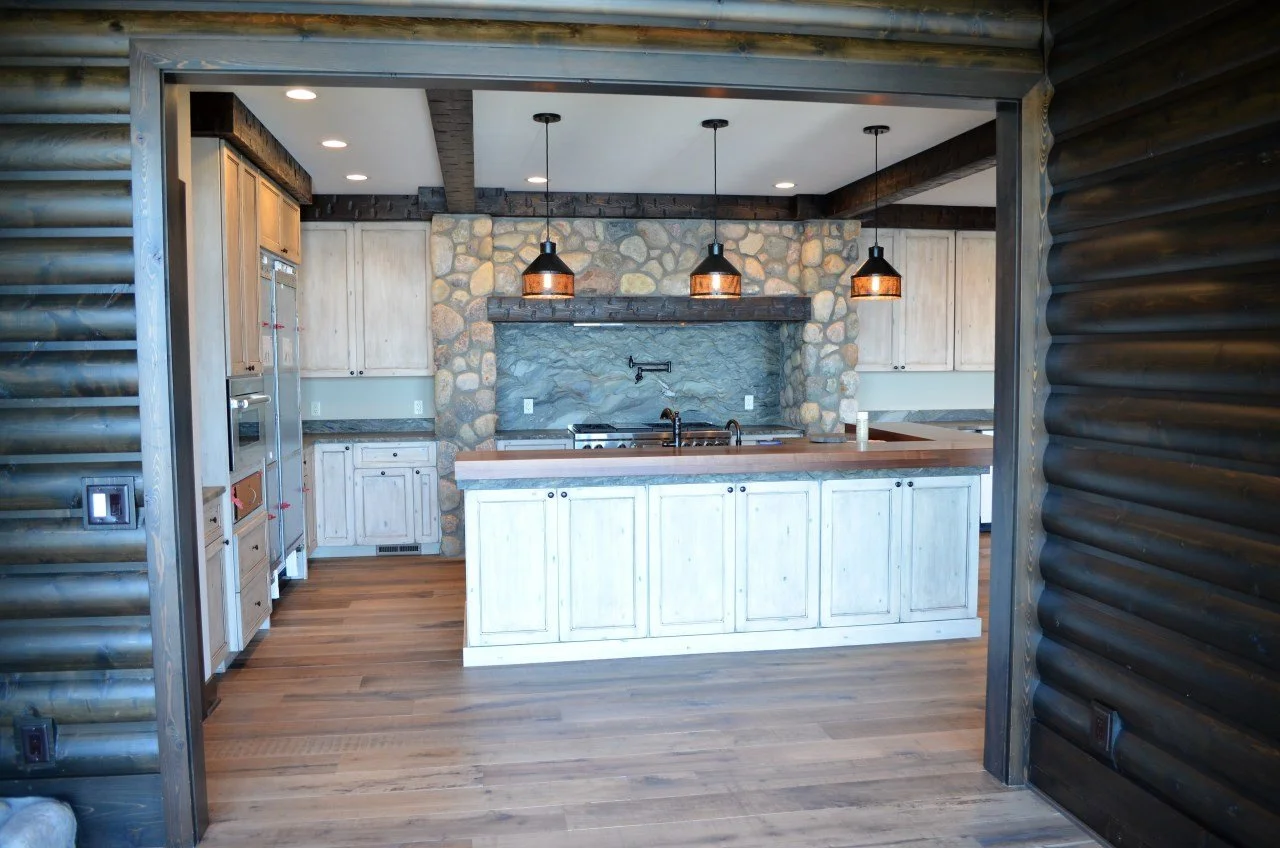 Kitchen with wooden cabinets, stone wall backsplash, wooden island, pendant lights, and wooden flooring in a rustic cabin style.
