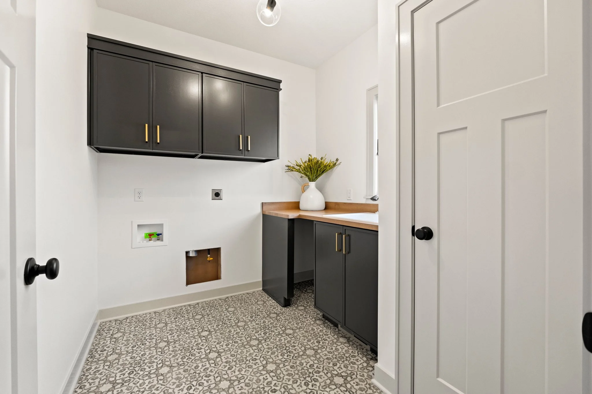 Laundry or utility room with white walls, black cabinets with gold handles, a beige countertop, a white vase with green plants, patterned tile flooring, and an open wall space with plumbing access and electrical outlets.