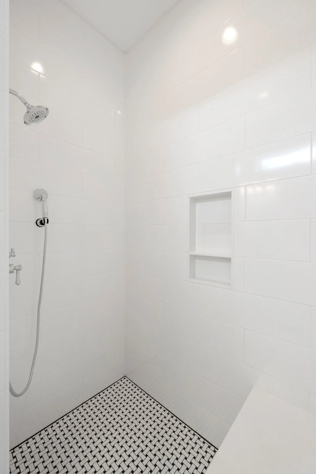 A minimalist, white-tiled shower with a built-in shelf, a rain showerhead, and a handheld shower.