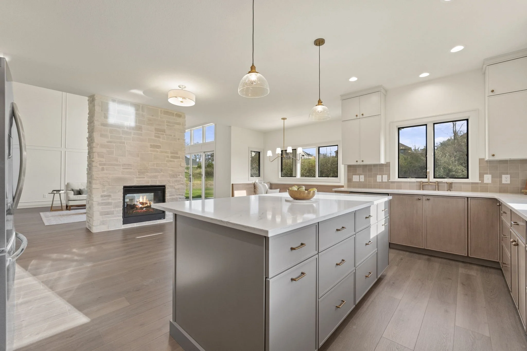 Bright, modern kitchen with white cabinets, an island with a white countertop, three pendant lights, large windows, a fireplace in the adjacent living area, and hardwood floors.