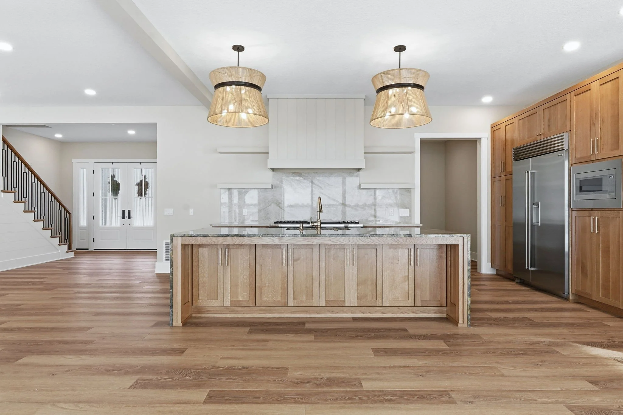 Modern kitchen with wooden cabinets, marble backsplash, pendant lights, stainless steel refrigerator, and an island with a sink.