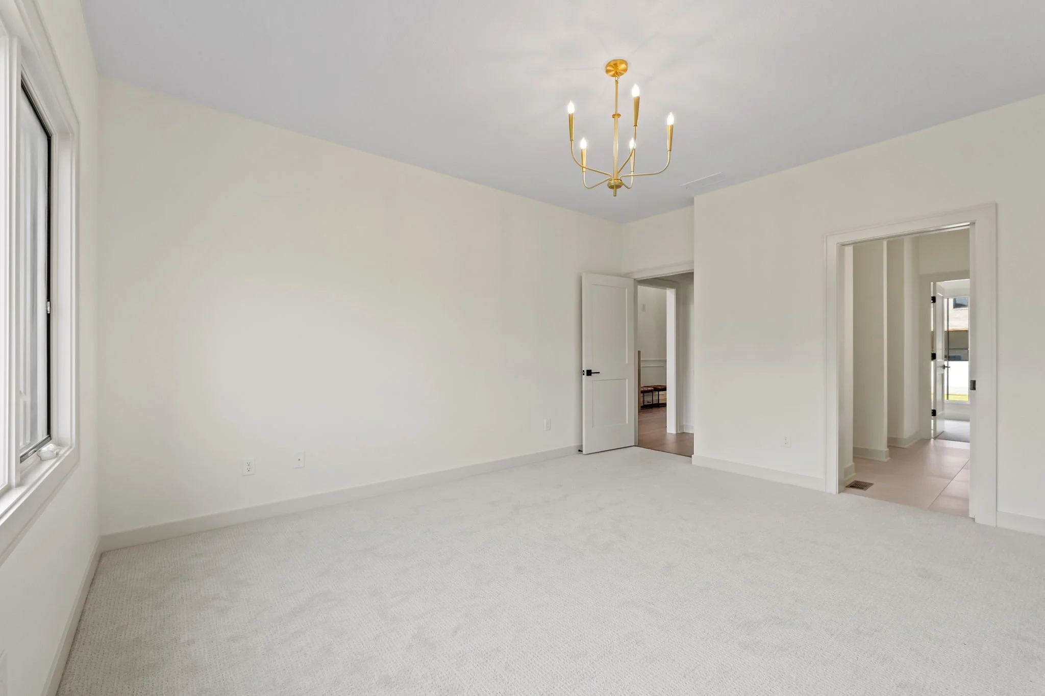 Empty room with white walls, white carpeting, a window on the left, and a modern chandelier hanging from the ceiling. Multiple doorways leading to other areas of the house.