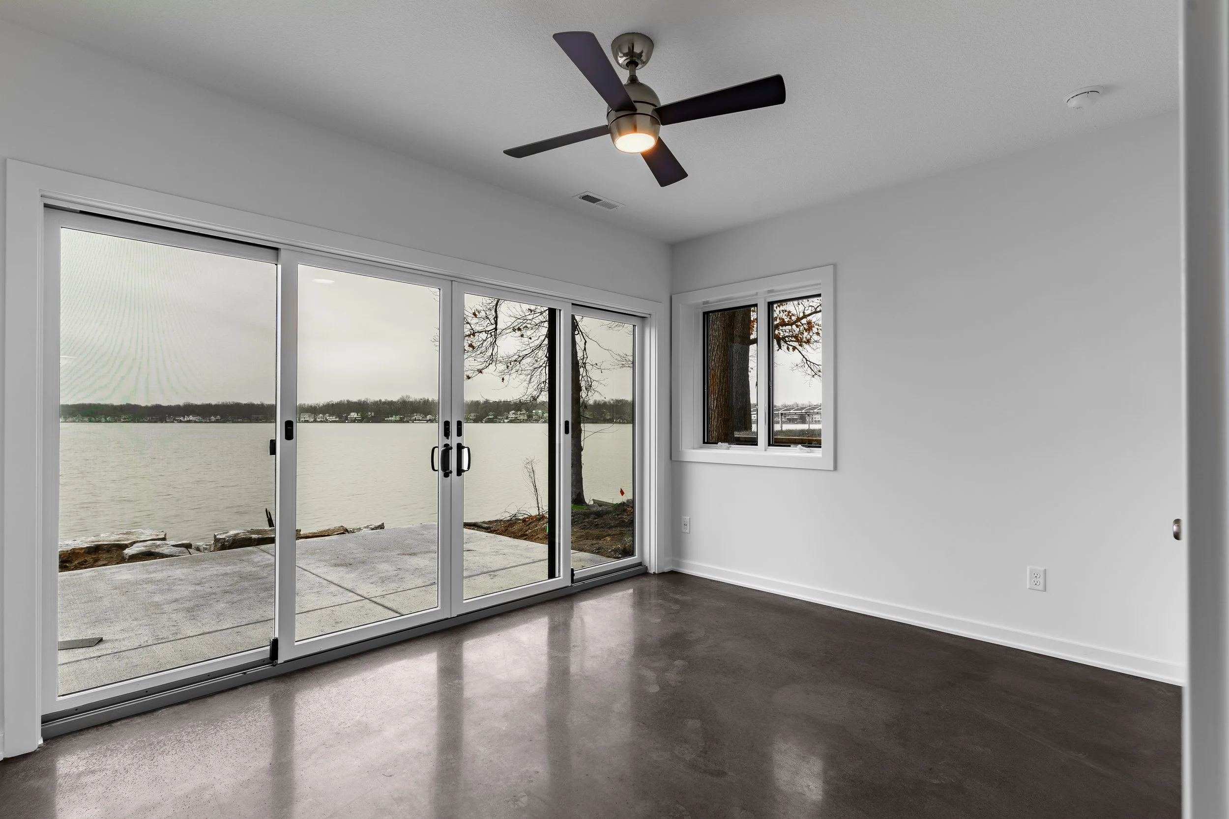 Empty room with white walls, brown floor, ceiling fan, large sliding glass door leading to a patio overlooking a lake, and a window showing trees outside.