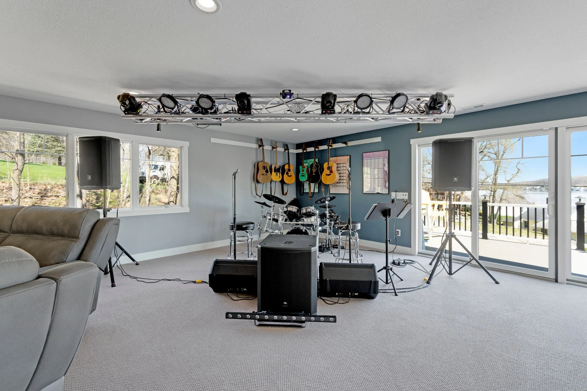 Music performance space with guitars on the wall, electronic drum set, speakers, microphones, and stage lighting, overlooking a waterfront view.