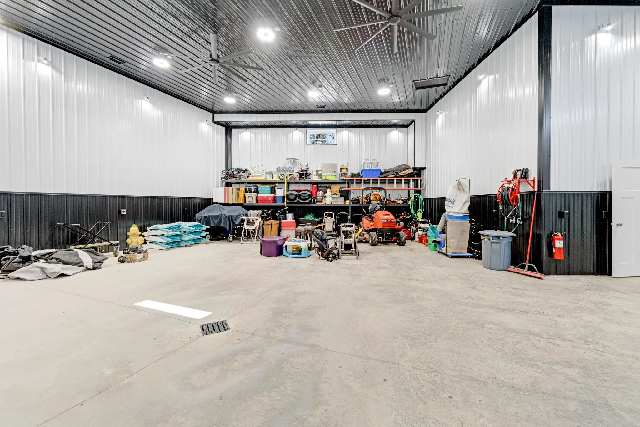 A spacious garage with a concrete floor, white and black paneled walls, and a black metal ceiling with ceiling fans and bright lighting. Various tools, lawn equipment, and storage items are organized on shelves and on the floor.
