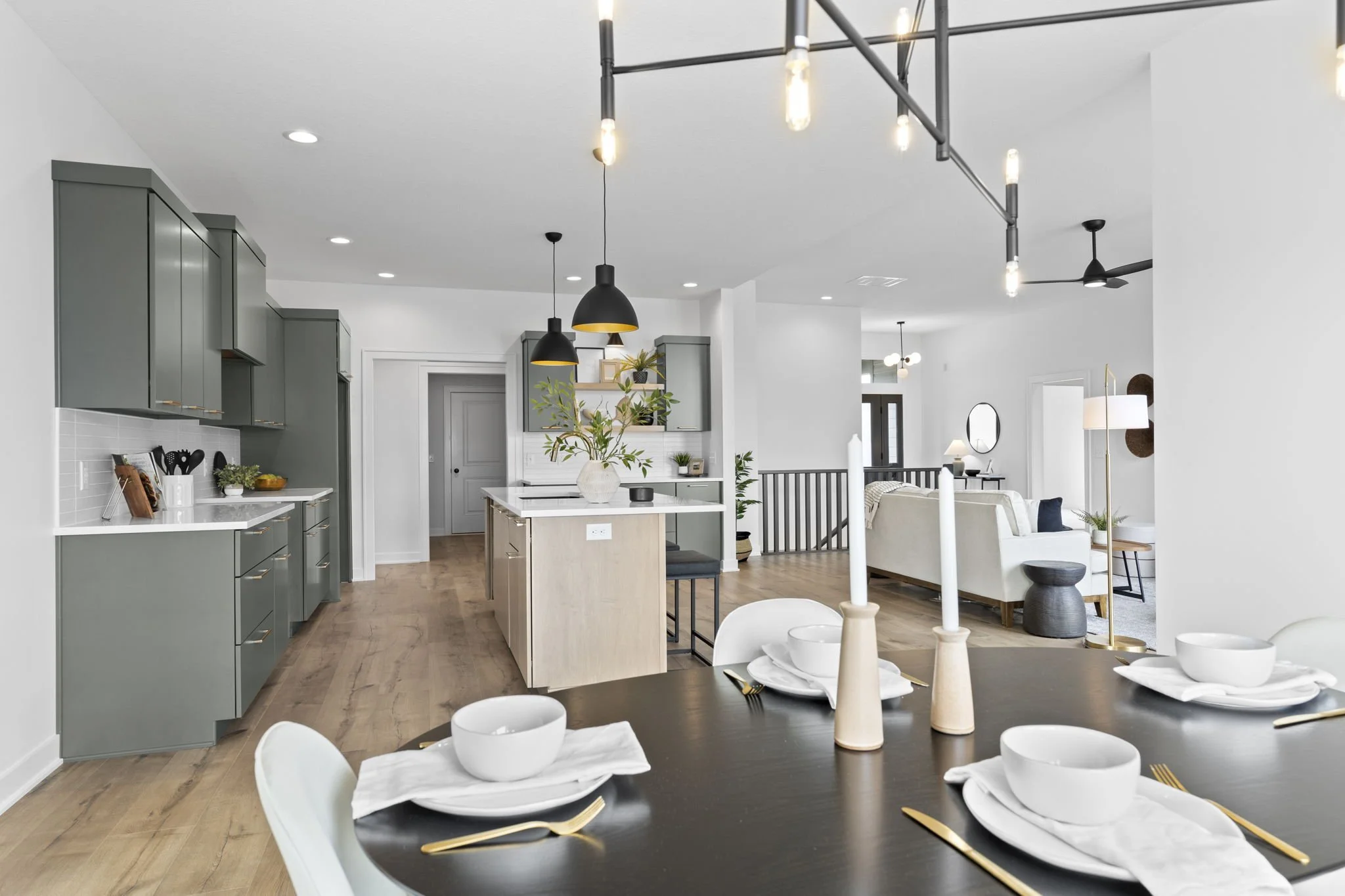 Open-concept kitchen and dining area with modern decor, sage green cabinetry, and a wooden kitchen island with black pendant lights, white walls, and minimalistic table setting with candles and bowls.