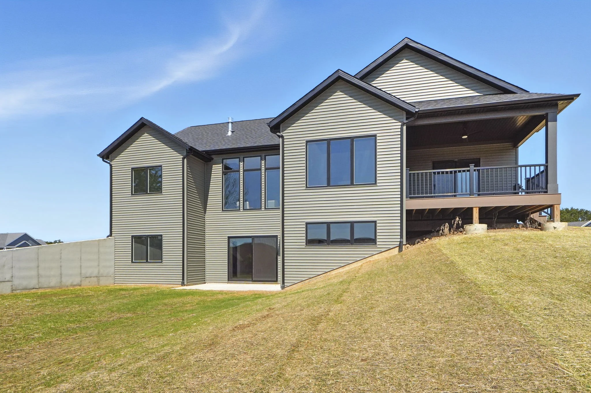 A modern, two-story house with beige siding, black window trims, and a dark roof, situated on a grassy hill with a clear blue sky in the background.