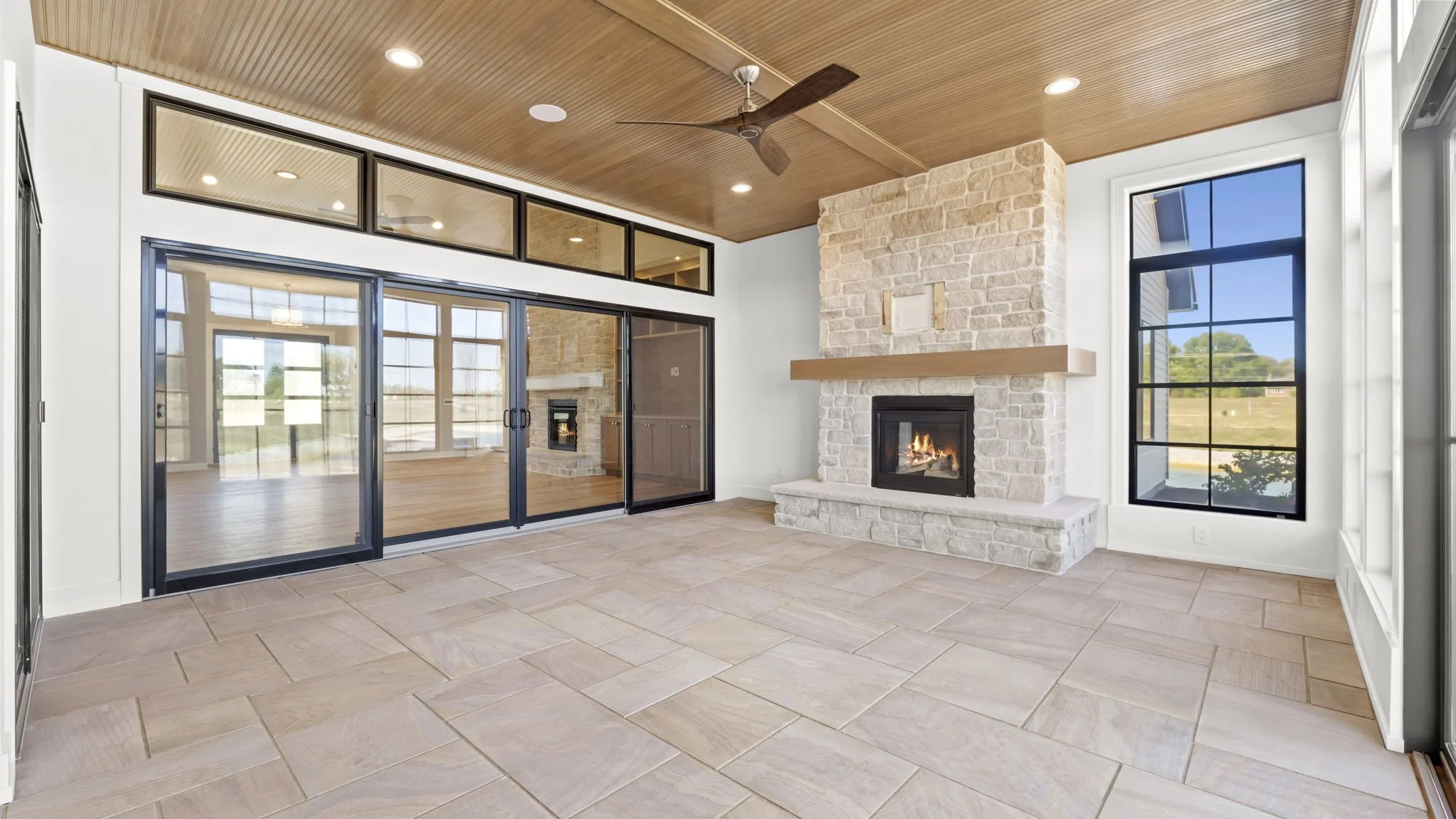 Interior of a modern room with a fireplace, large windows, and sliding glass doors leading to an outdoor area, with a wood-panel ceiling and tile flooring.