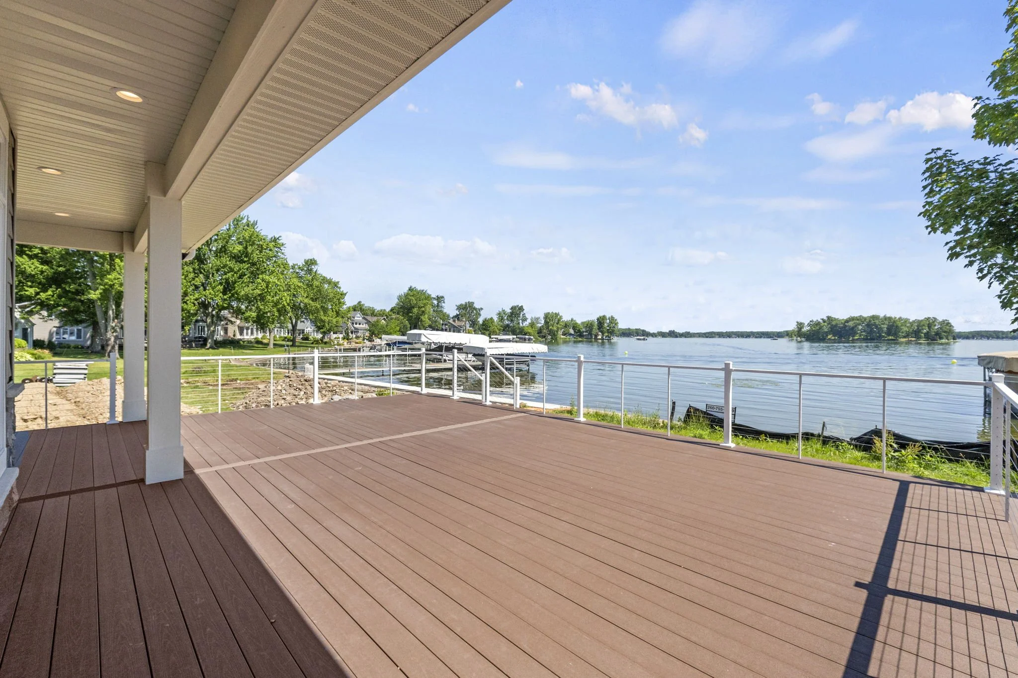 View of a large wooden deck overlooking a river with a retaining wall, trees, and small islands in the distance under a blue sky with some clouds.
