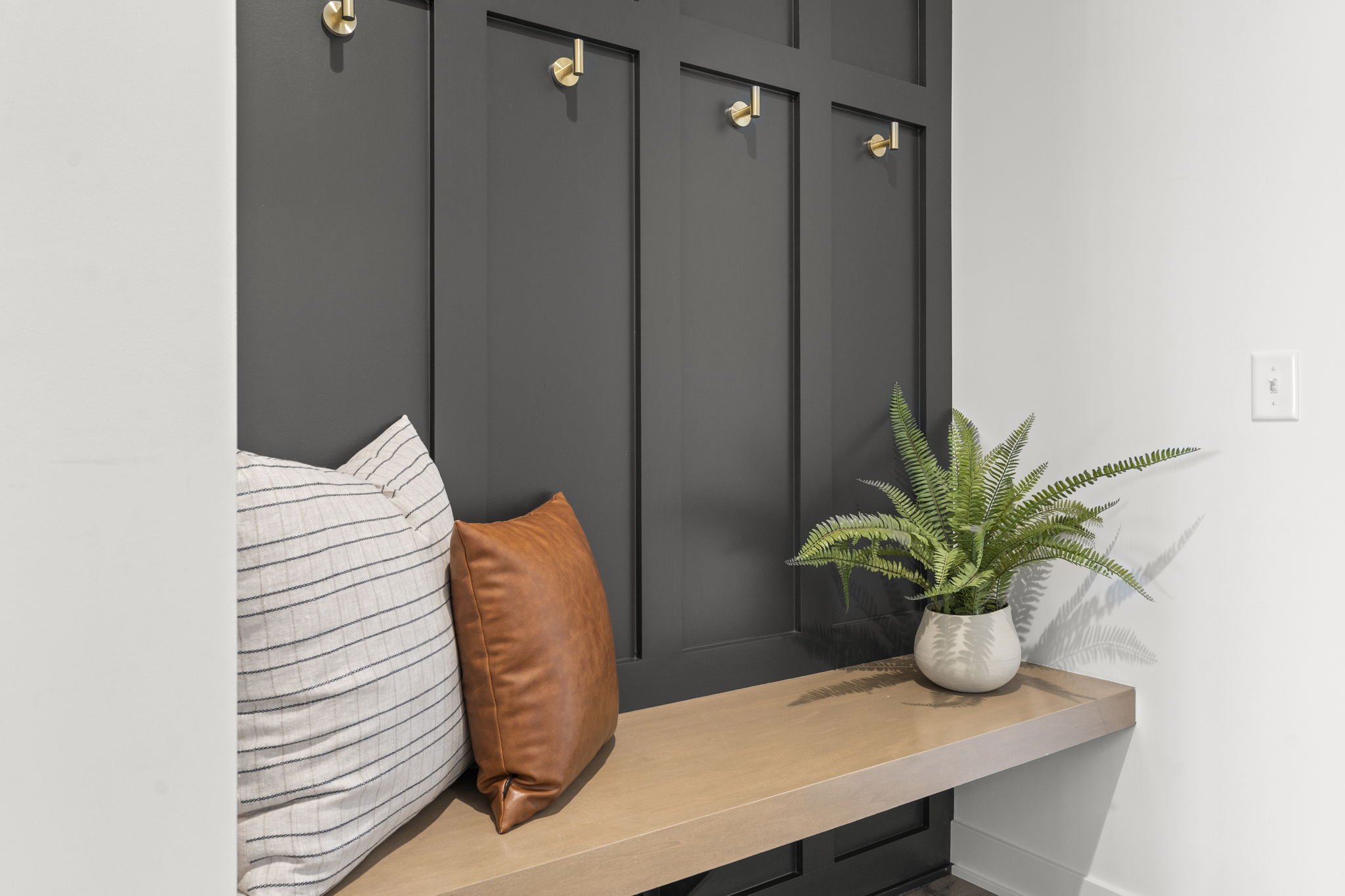 Close-up of a wooden bench with striped and leather pillows, a potted fern, against a black wall with gold hooks.