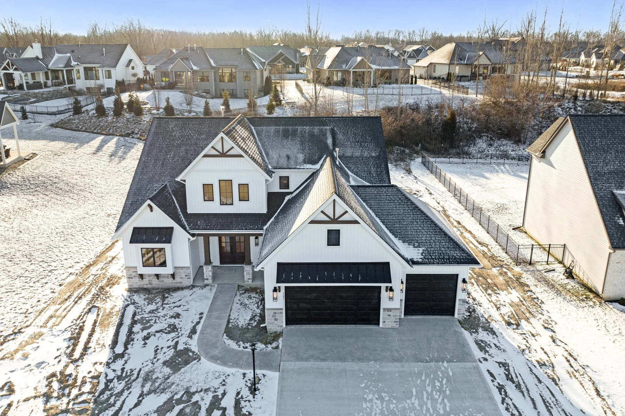 A large modern two-story house with white siding and black roof, surrounded by snow, with a concrete driveway and walkway, in a suburban neighborhood with other houses in the background.