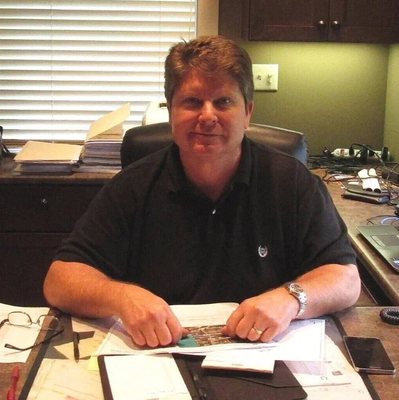 A man sitting at a cluttered desk in an office, smiling at the camera.