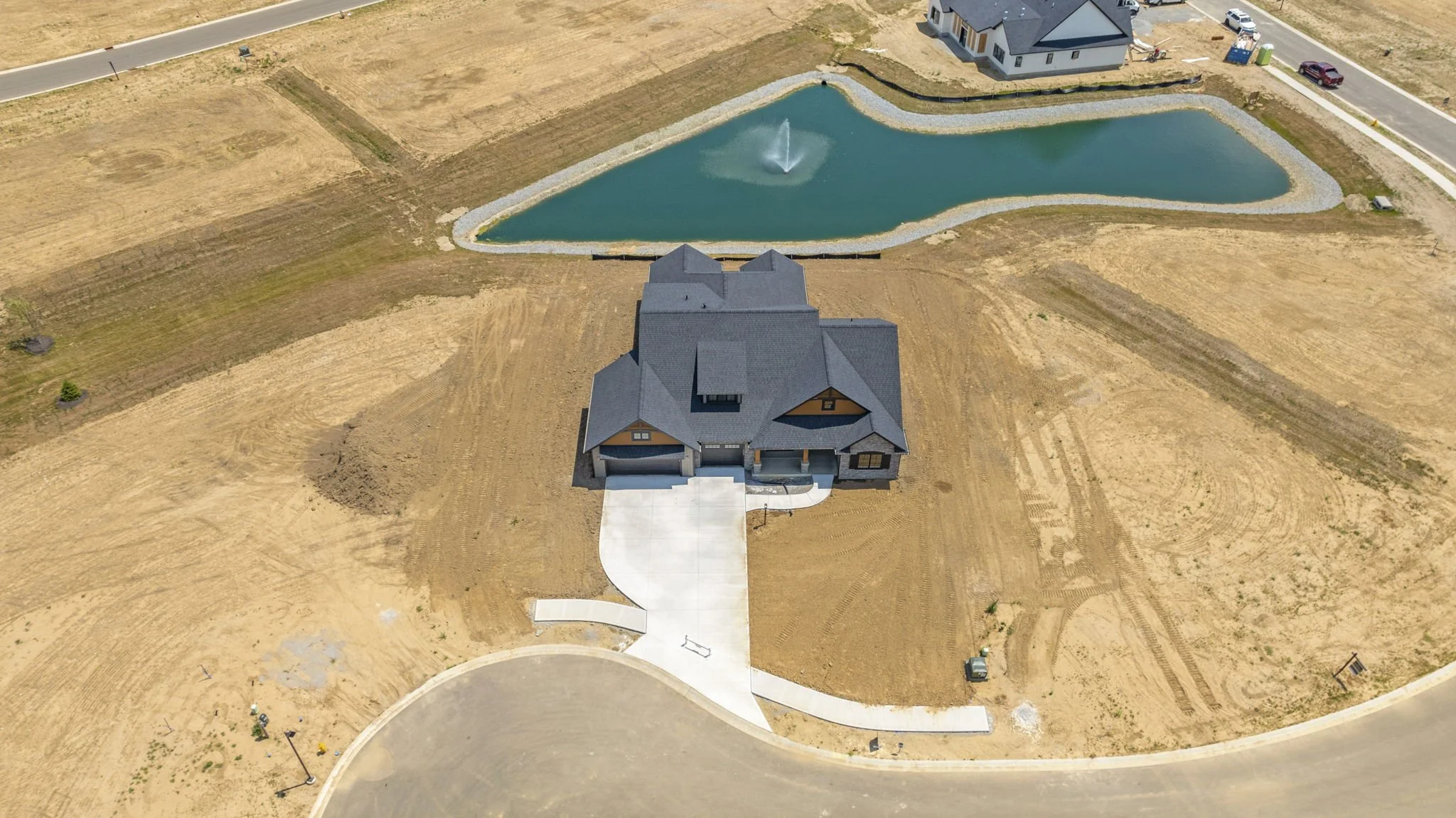 Aerial view of a house under construction with a dark gray roof, a curved driveway, and a pond with a fountain in the backyard. The surrounding land is mostly bare dirt.