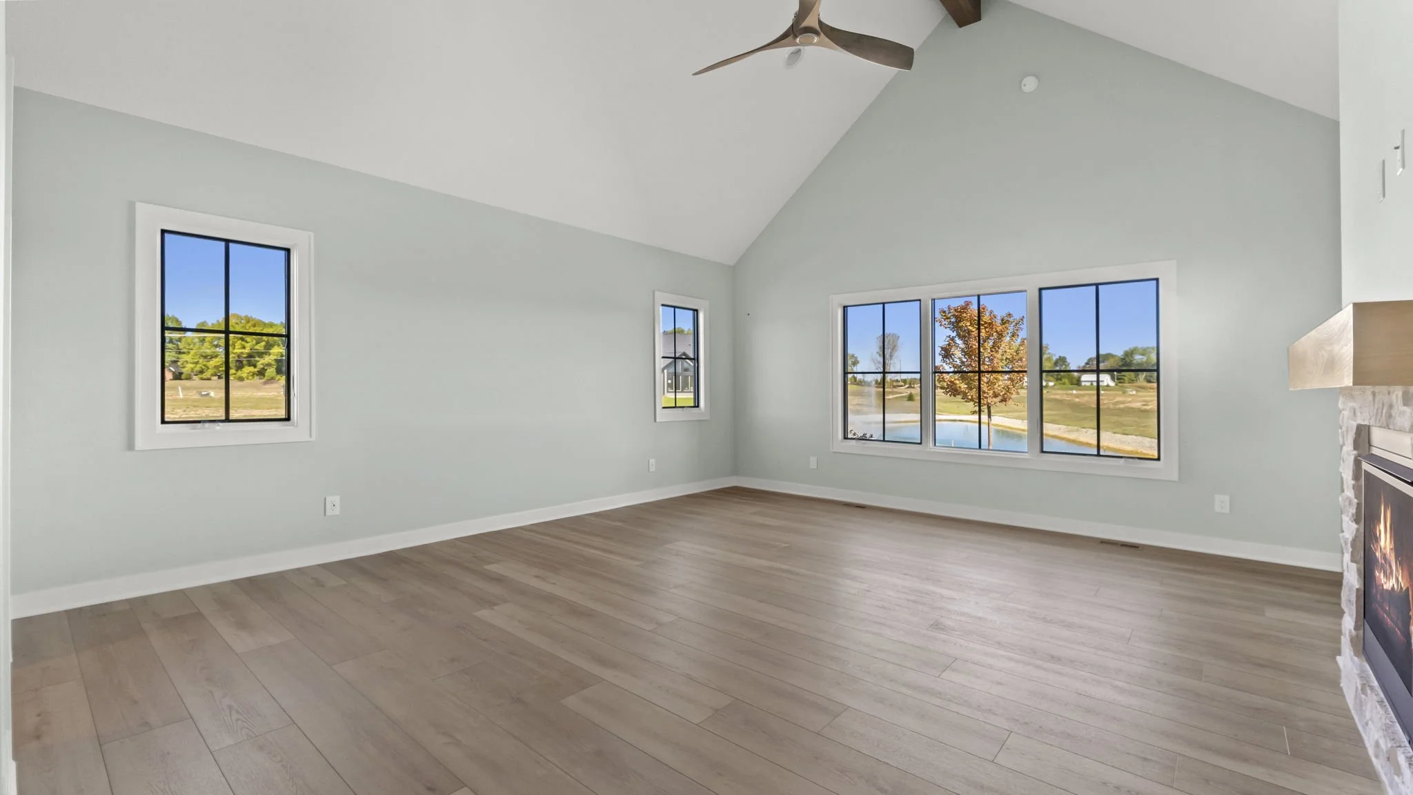Empty living room with light-colored wood flooring, pale blue walls, three large windows, a fireplace with a stone surround on the right, and a ceiling fan.