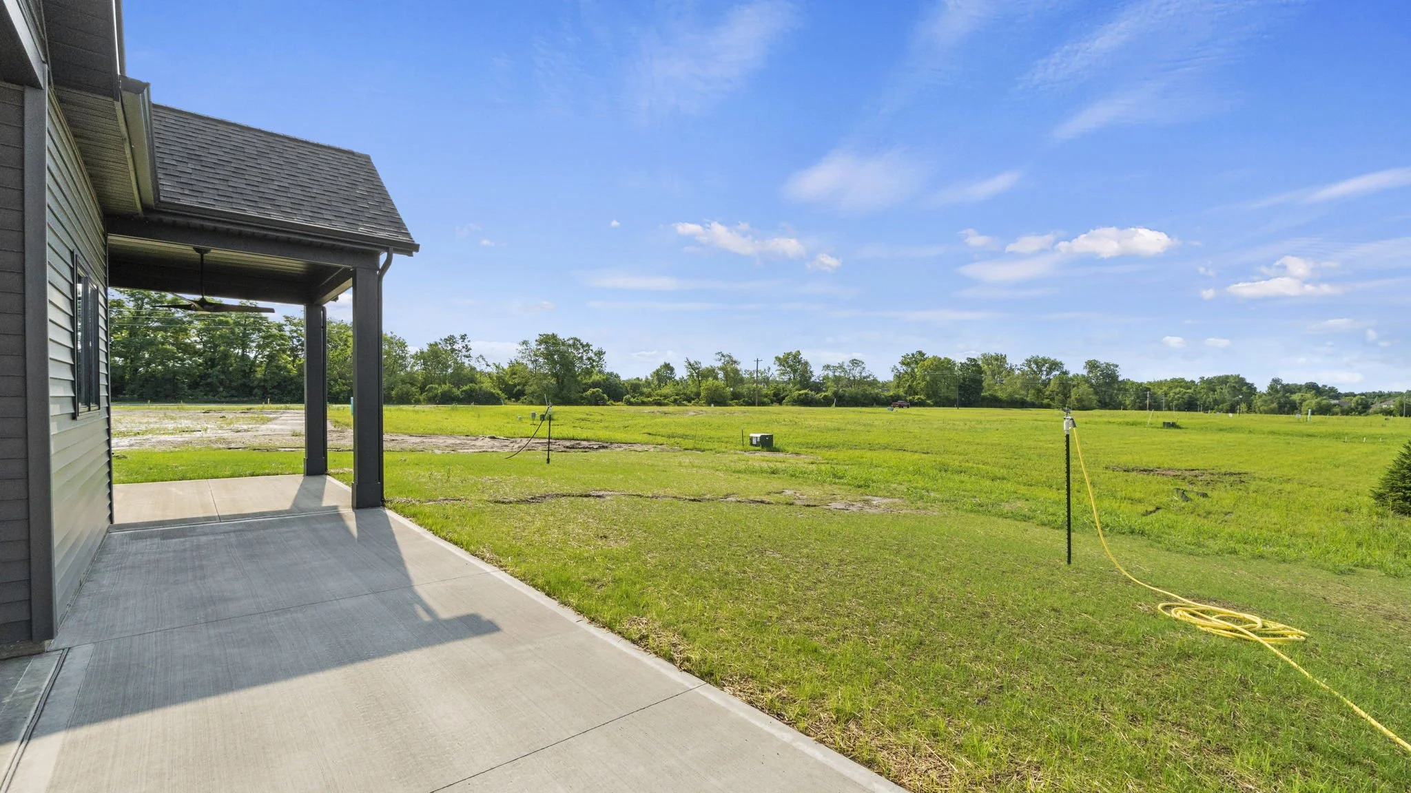 View of a backyard with a concrete patio, extending into a grassy field with a few small trees and clear blue sky.