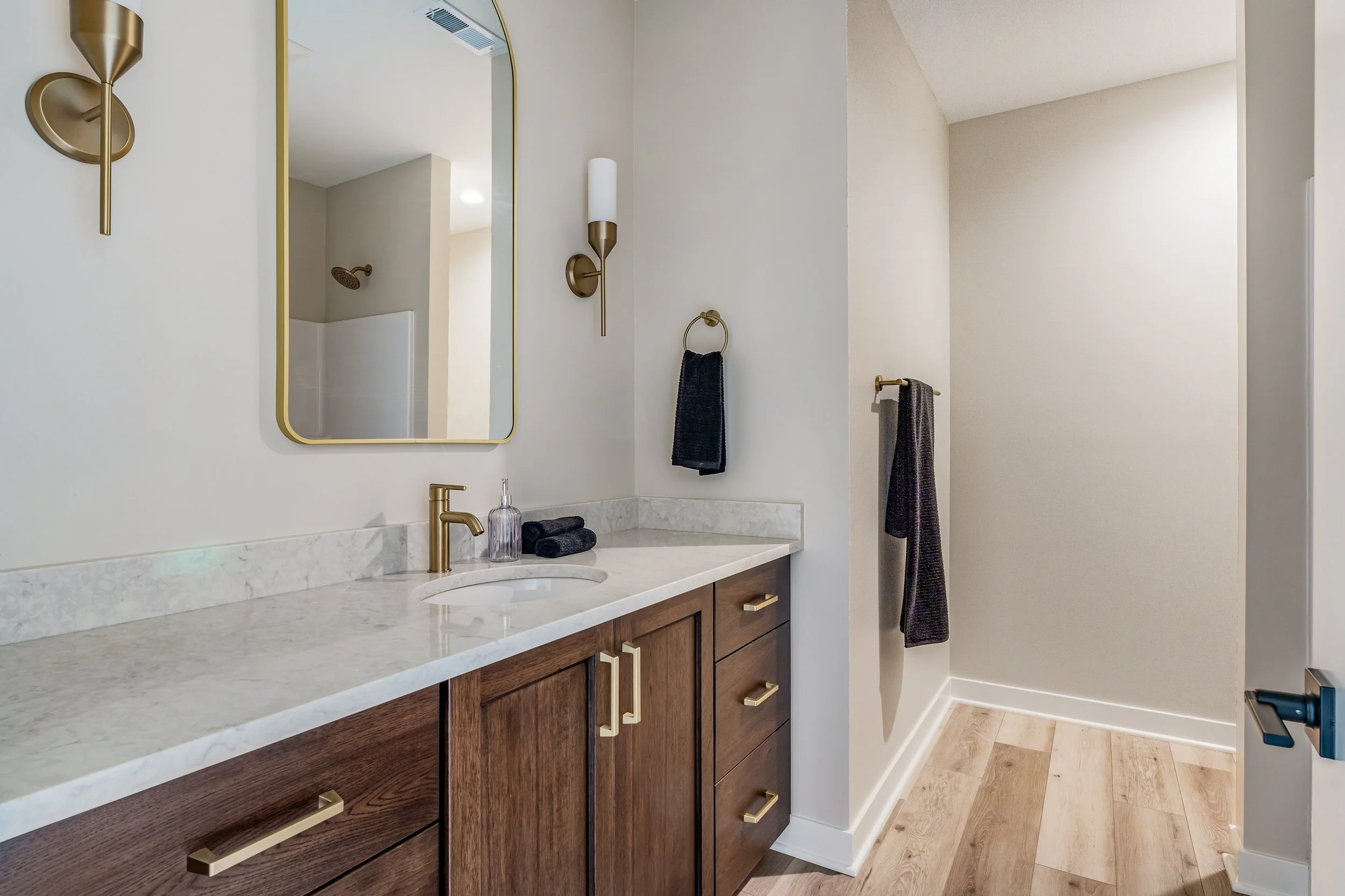 Bathroom vanity with a marble countertop, a gold faucet, and wooden cabinets with gold handles. A large mirror is mounted above, flanked by two gold wall sconces with white shades. Two dark towels are on the countertop, and two dark towels hang on wa