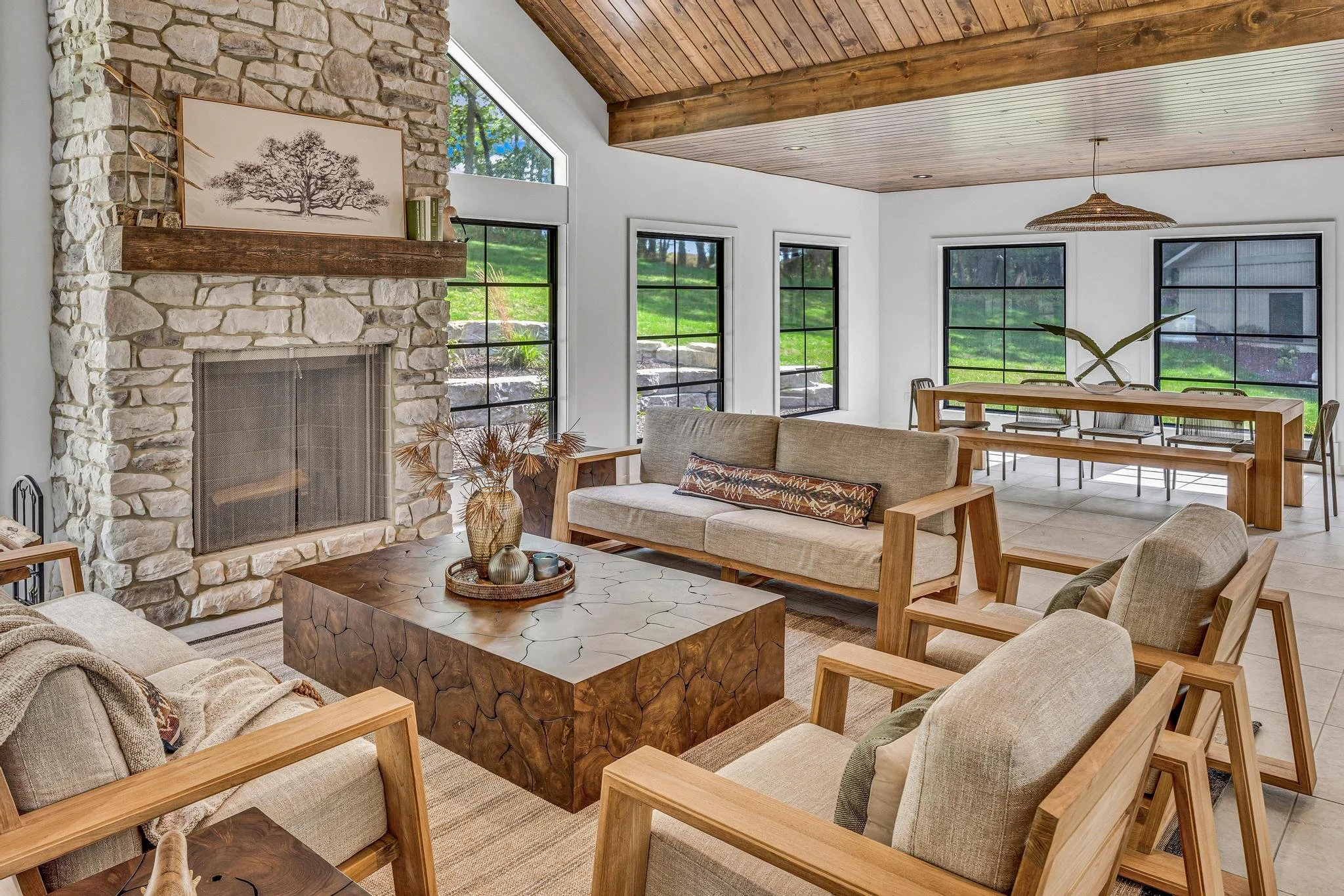 Living room with stone fireplace, large windows, wooden ceiling, beige sofas, armchairs, a wooden table, and a dining area with a long wooden table and chairs, decorated with black metal frames and greenery.