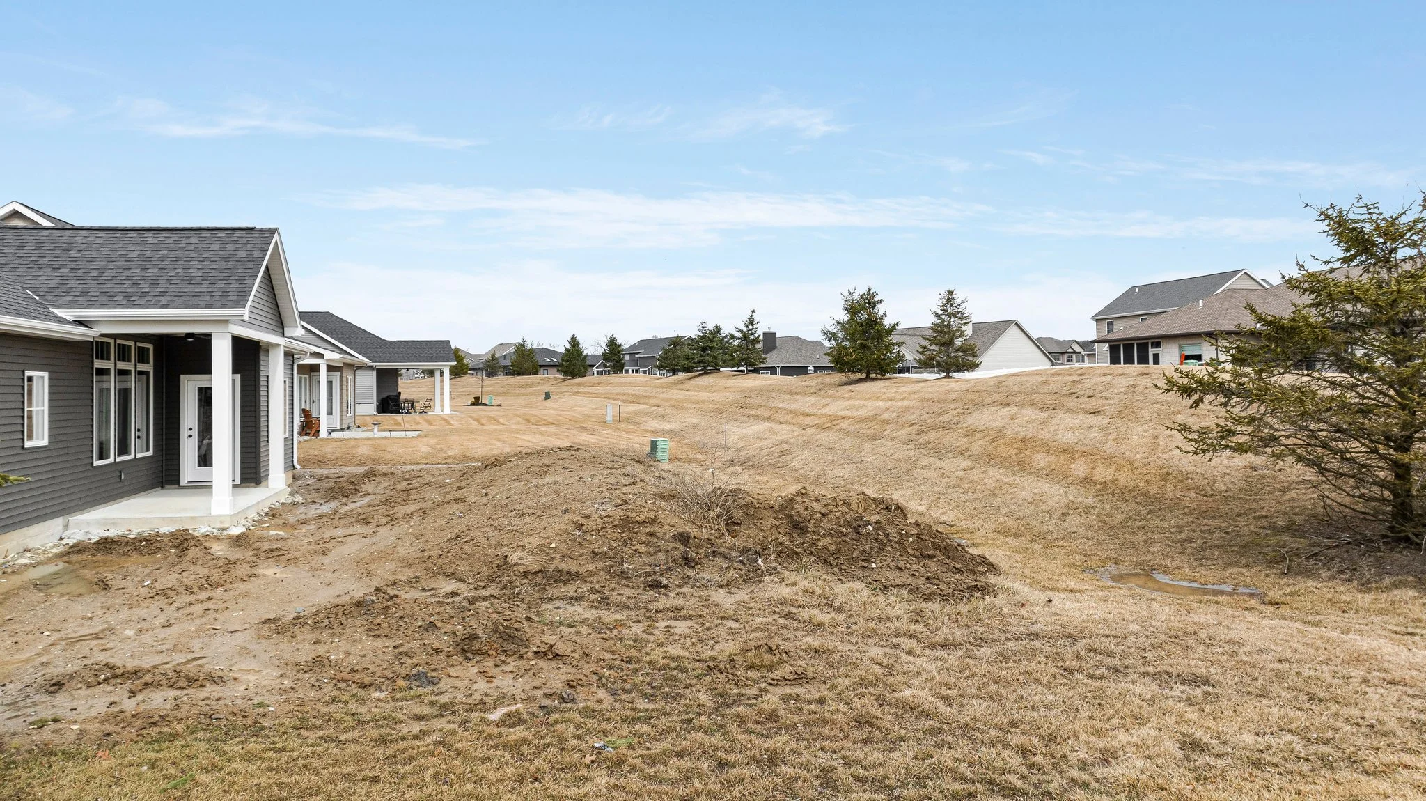 Side yard of a residential neighborhood with grass, dirt, and several houses in the background under a partly cloudy sky.