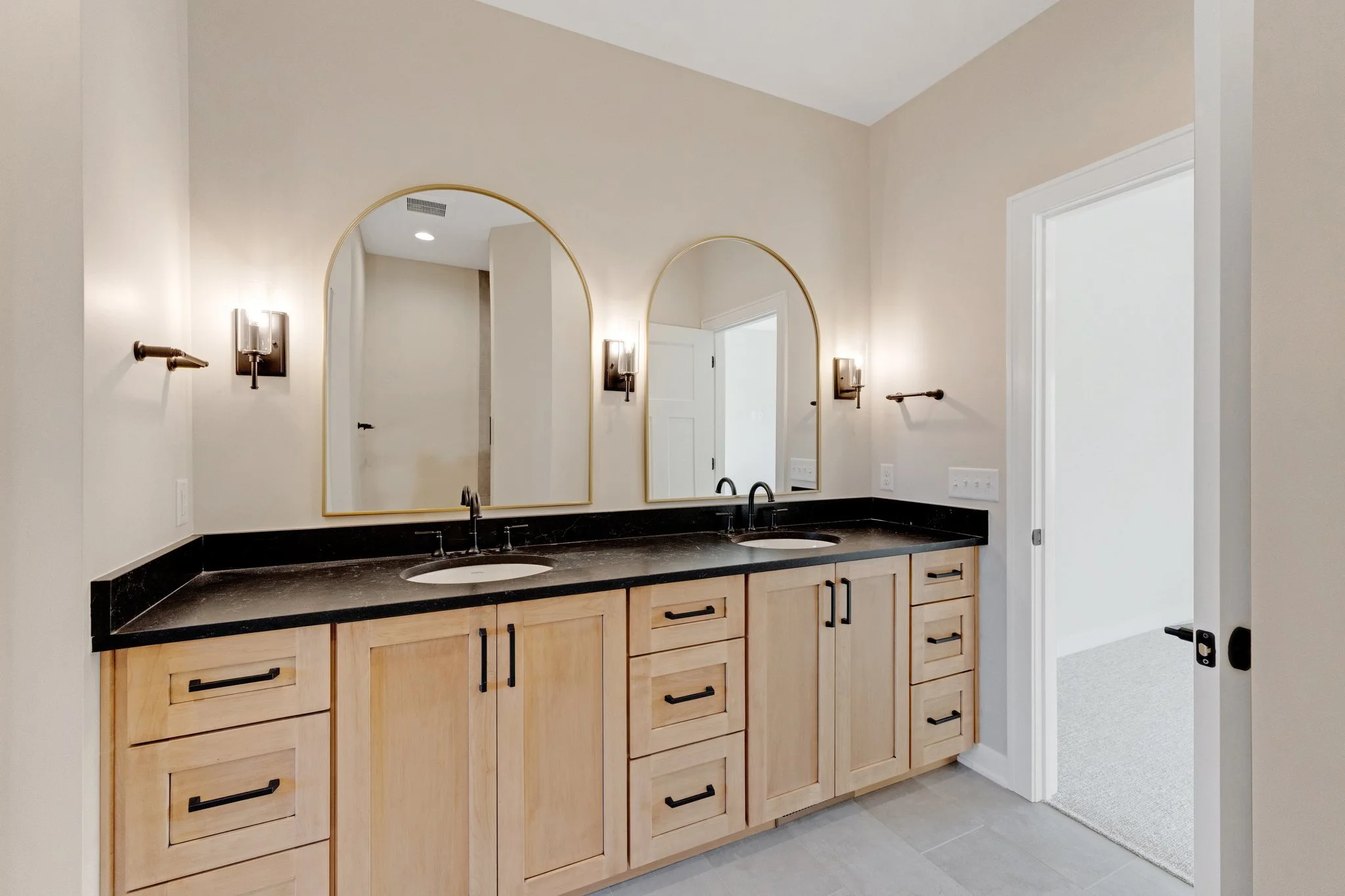 Modern bathroom with double vanity featuring light wood cabinets, black countertop, two oval mirrors, and wall-mounted lighting fixtures.