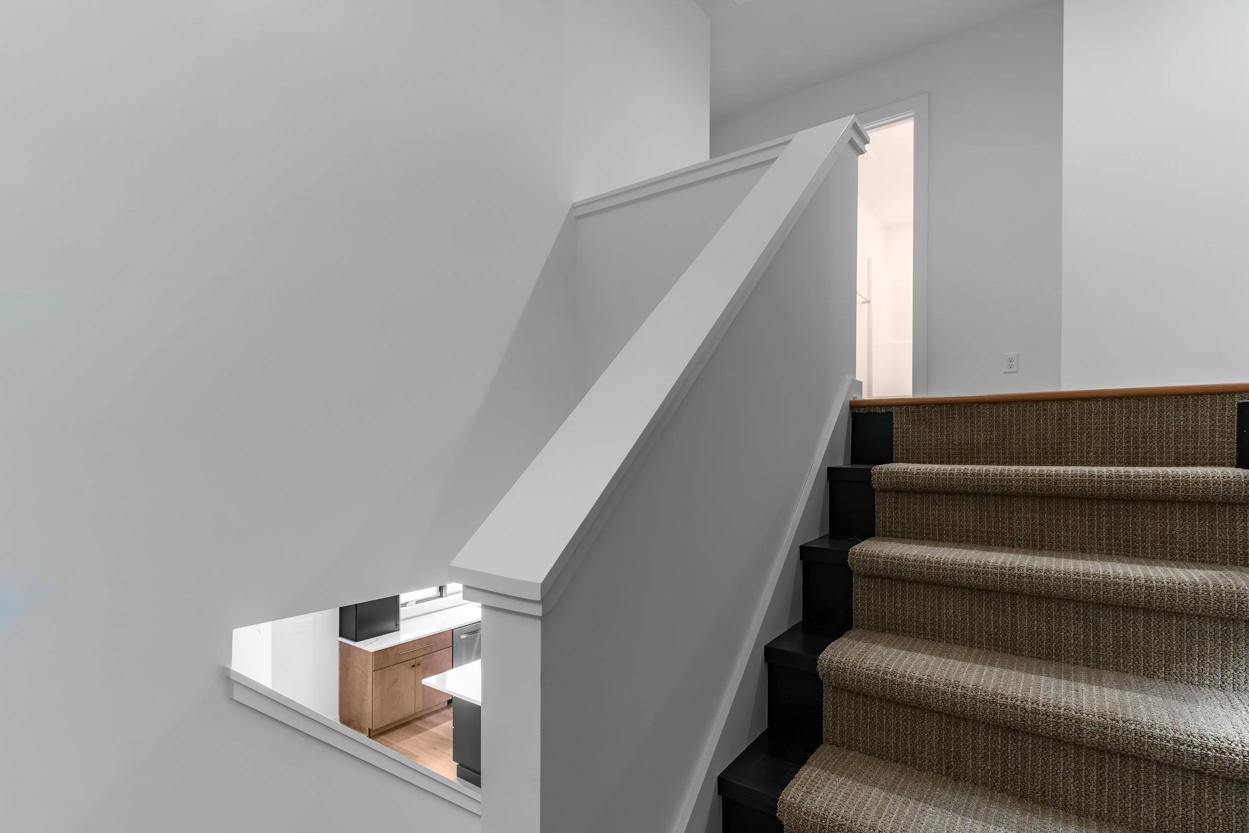 Interior view of a staircase with dark wood steps, a beige patterned carpet runner, and a white painted railing, leading to a bright doorway, with a window on the wall showing a partial view of a kitchen area.