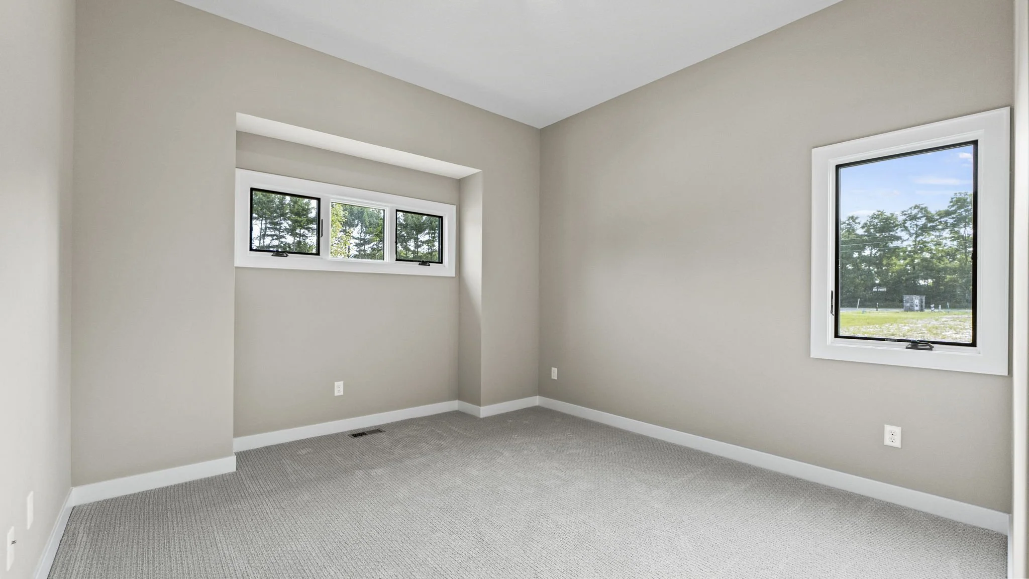 Empty room with beige walls, gray carpeting, two small horizontal windows, and a large vertical window, with trees visible outside.