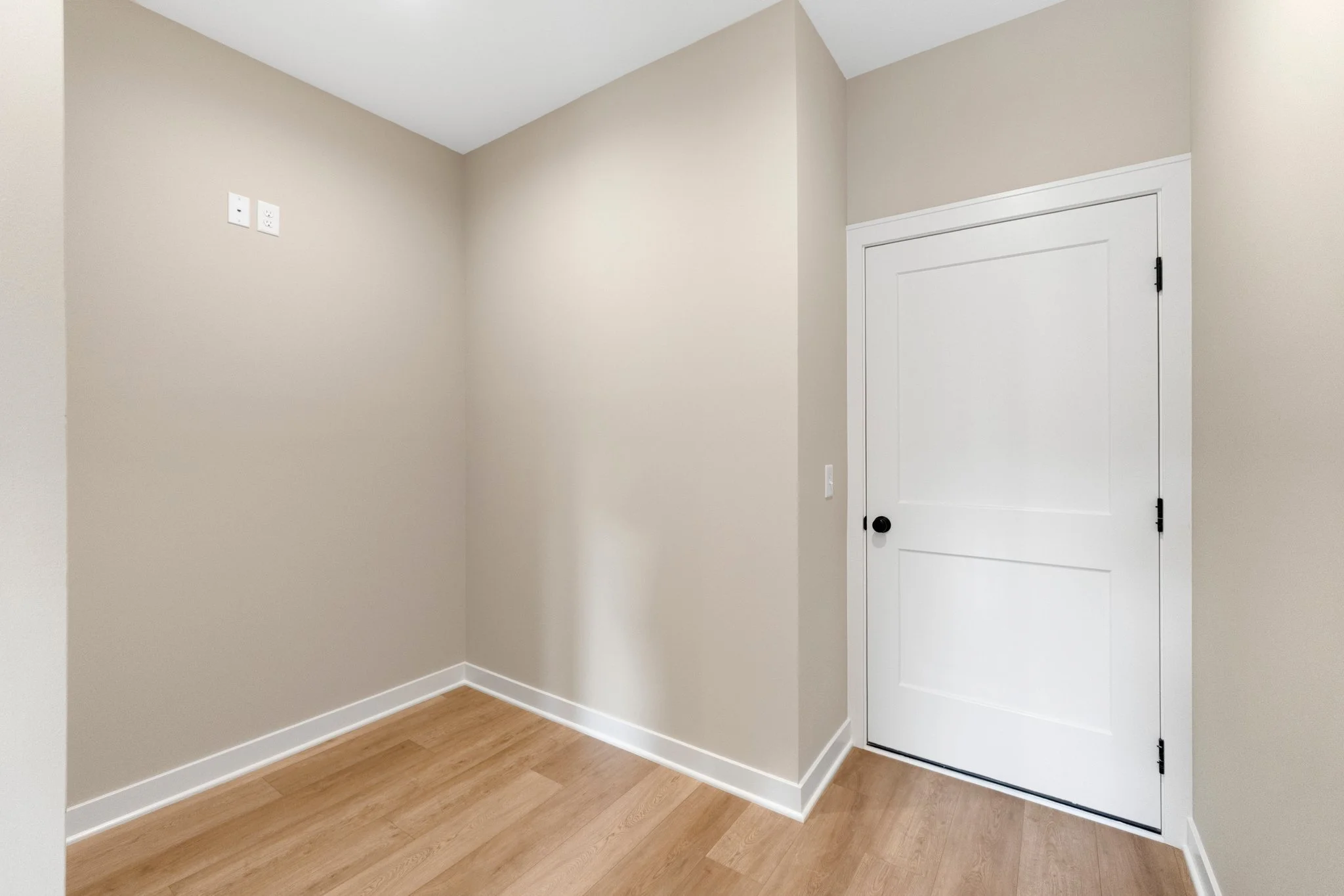 Empty room with beige walls, white trim, a white door with black hardware, and light wood flooring.