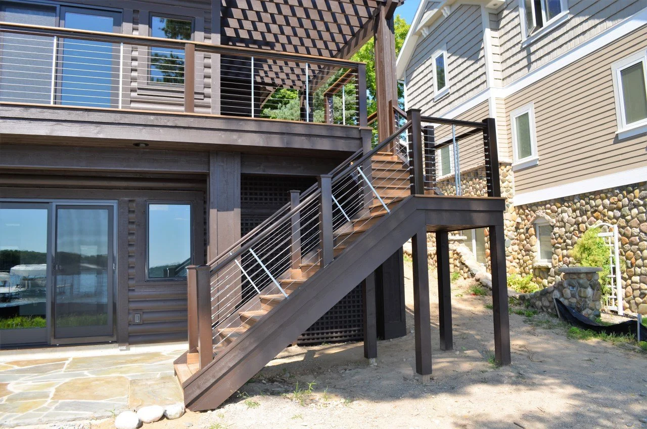 Exterior view of a house with a staircase leading to an upper deck. The house has beige siding and a stone foundation on one side. The deck has wooden planks and modern railing with metal cables. The area around the house is landscaped with some dirt