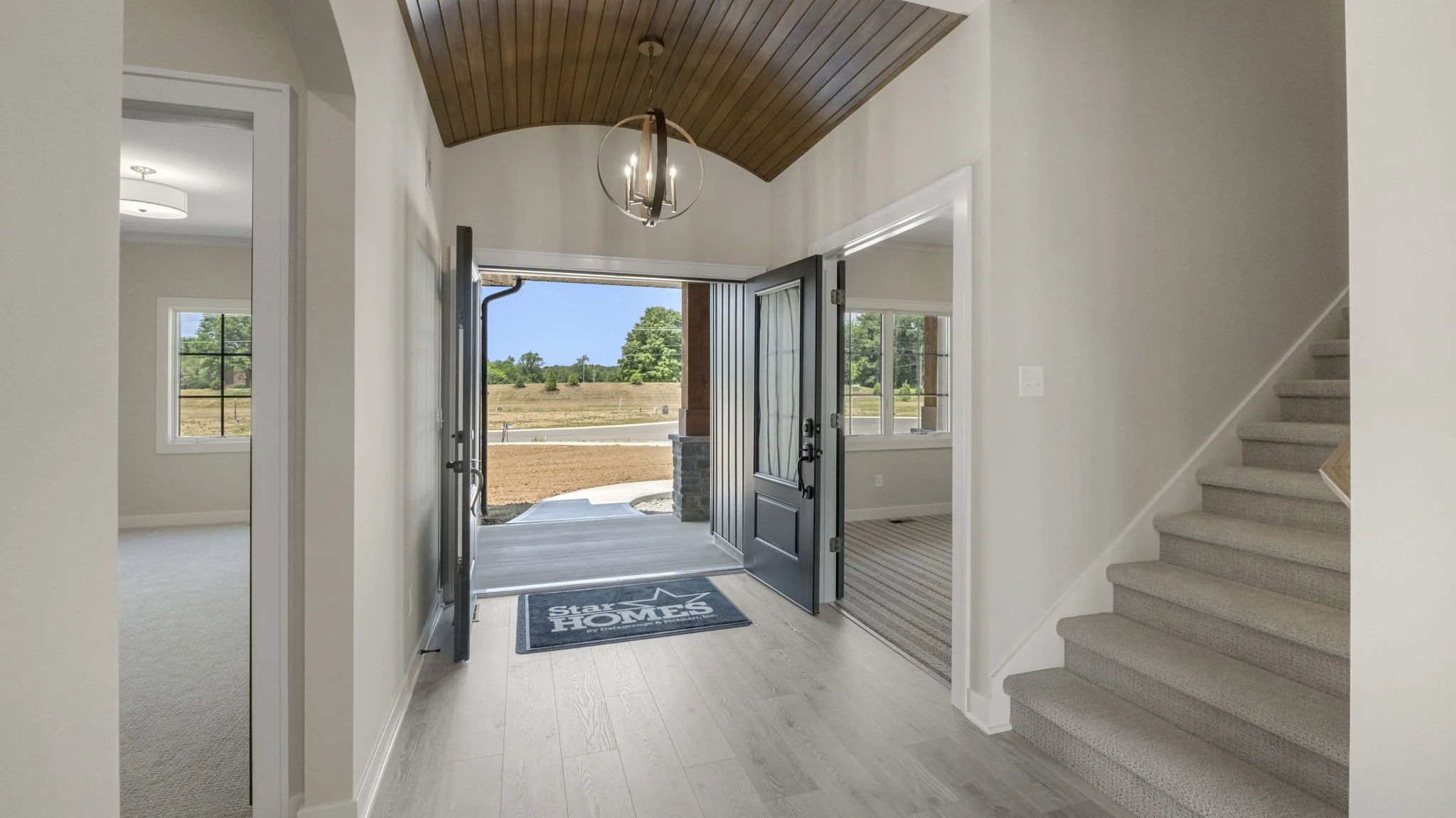 Entryway of a house with open front door leading outside to a partially landscaped yard, featuring a staircase on the right, and a hanging chandelier on a wooden ceiling.