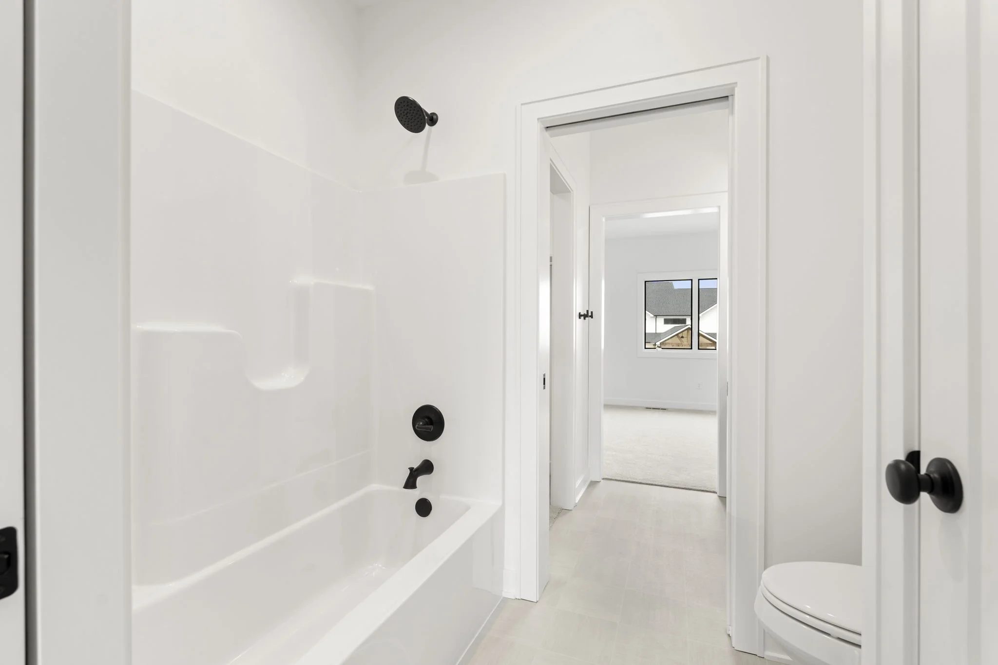 Modern white bathroom with bathtub, black fixtures, and a small window in an adjacent room.