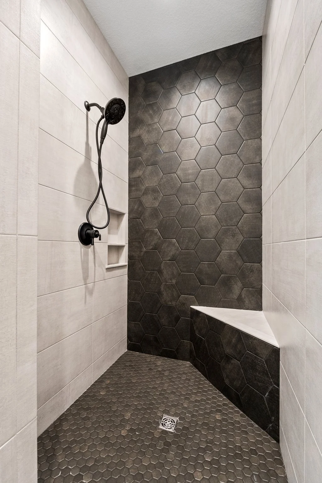 Modern shower with large beige tiles on the walls, black hexagonal tiles on a side wall, and small matching hexagonal tiles on the floor. A built-in shelf on the wall and a drain on the floor.