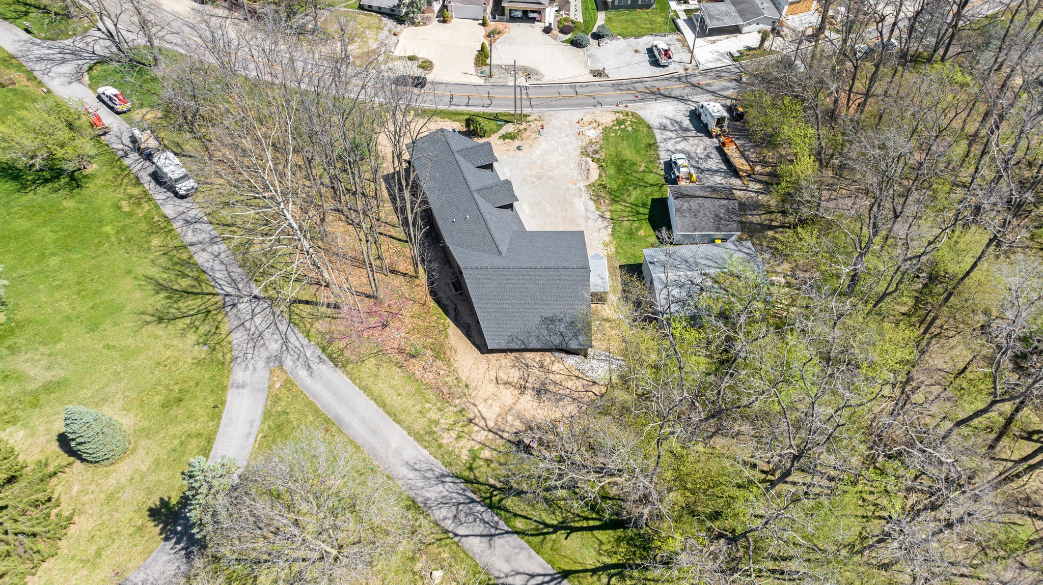 An aerial view of a house surrounded by trees with some leafless, a gravel driveway, adjacent grassy areas, and neighboring houses with paved roads, parked cars, and construction vehicles nearby.