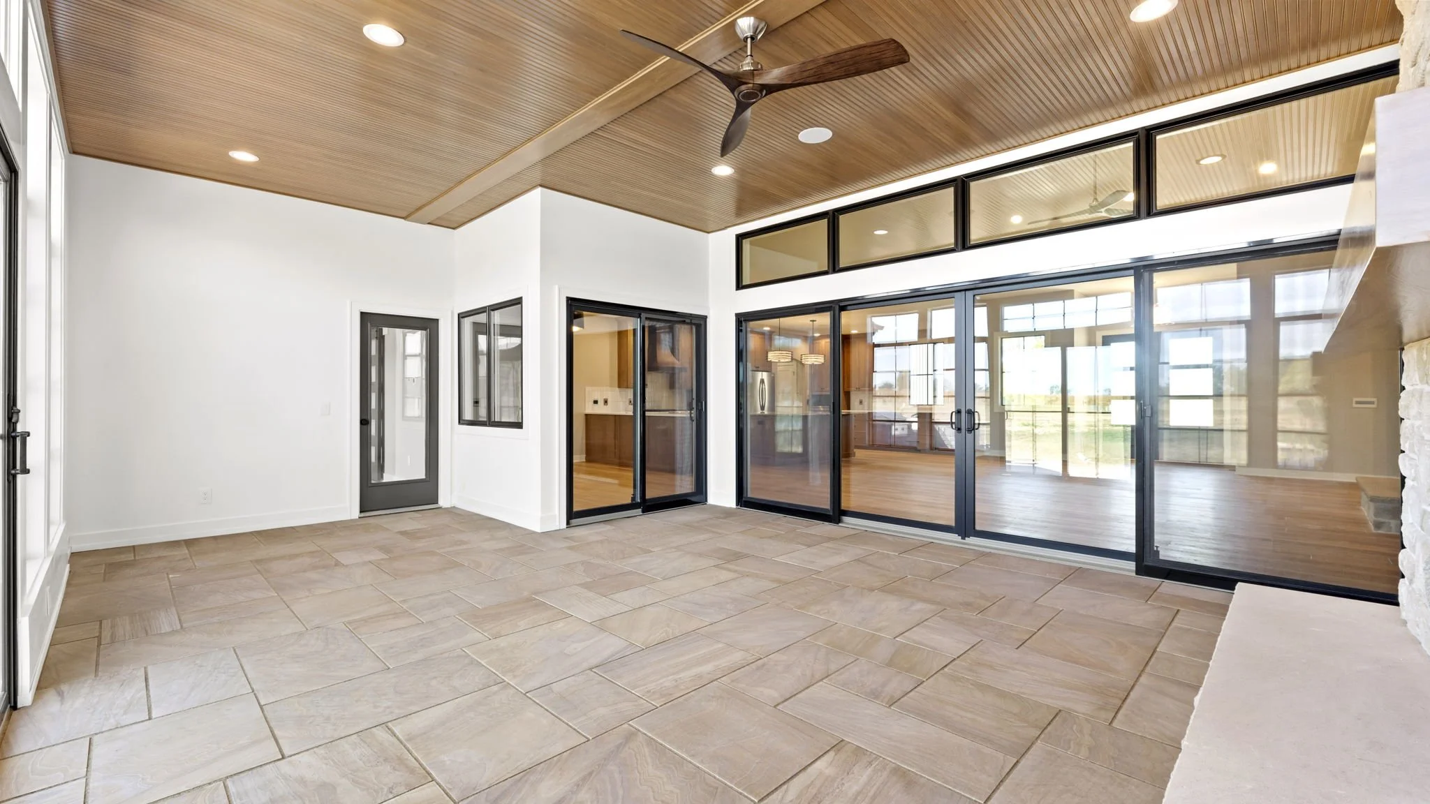 Empty sunroom with large tiled floor, white walls, wood-paneled ceiling, ceiling fan, and large glass sliding doors opening to a room with hardwood floors and windows.