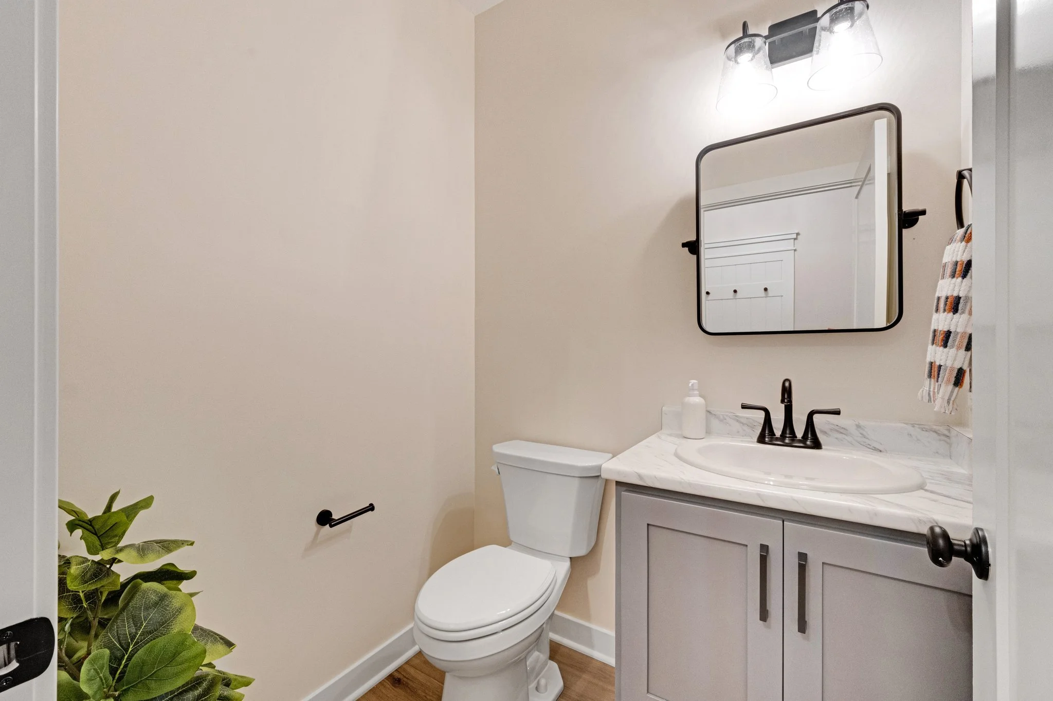 A small bathroom with a toilet, a light gray vanity with a marble countertop, a rectangular mirror, a black faucet, and a wall-mounted light fixture with clear glass shades. There is a towel hanging on a hook and a potted plant in the corner.
